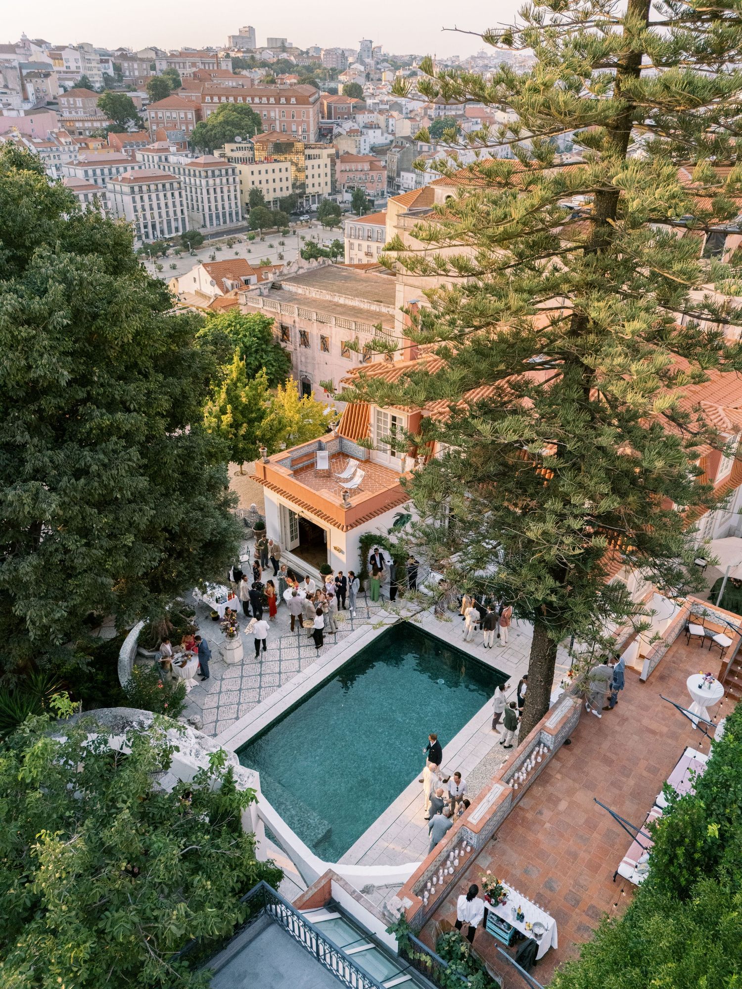 Aerial view of an elegant outdoor pool party at a hillside venue overlooking a European city at dusk.