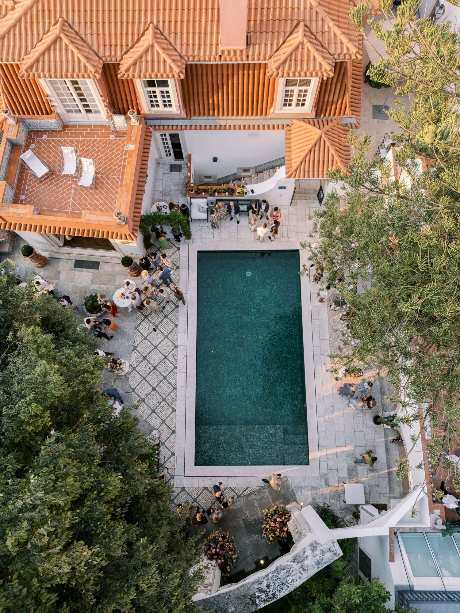 Aerial view of a luxury villa with orange tiled roof, rectangular pool, and lush green trees surrounding a courtyard.