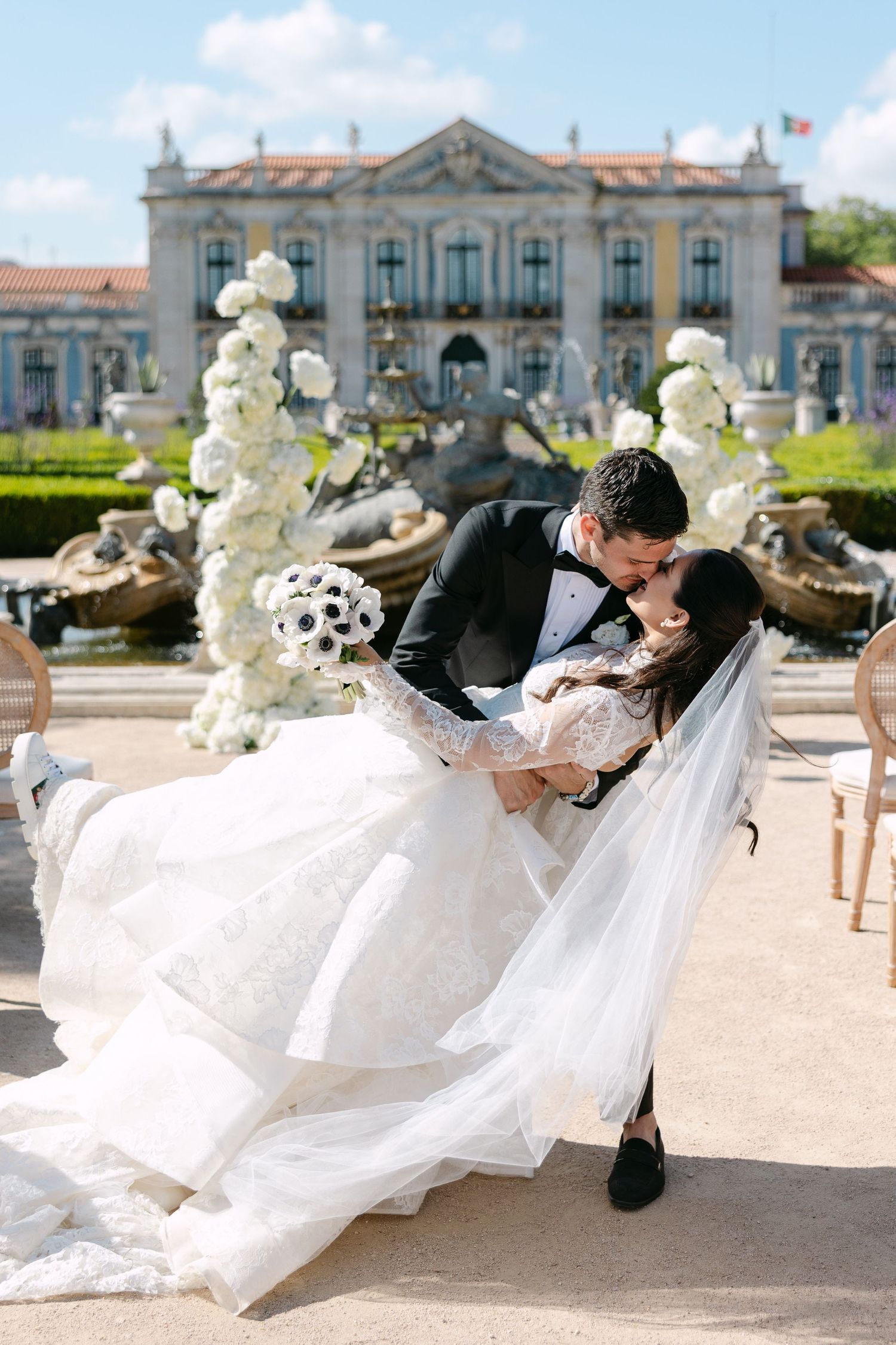 Groom dips bride for a kiss in front of an elegant palace fountain adorned with white floral arrangements.