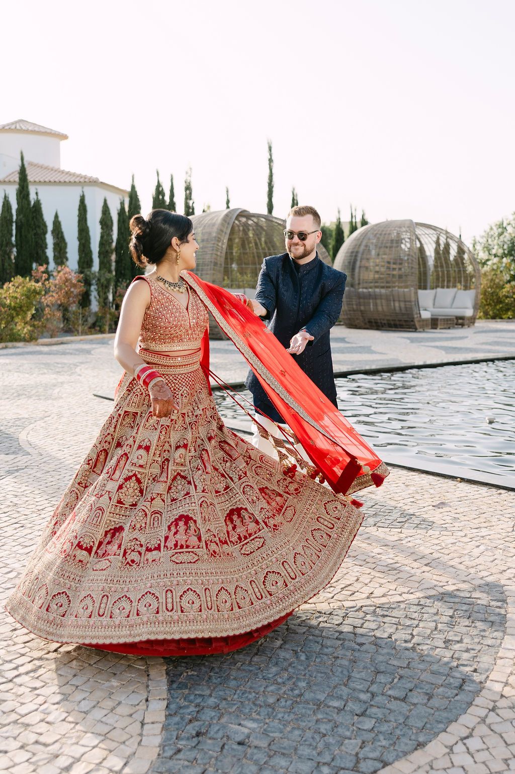 Beautiful red and gold embroidered lehenga twirls on cobblestone courtyard with cypress trees in background.