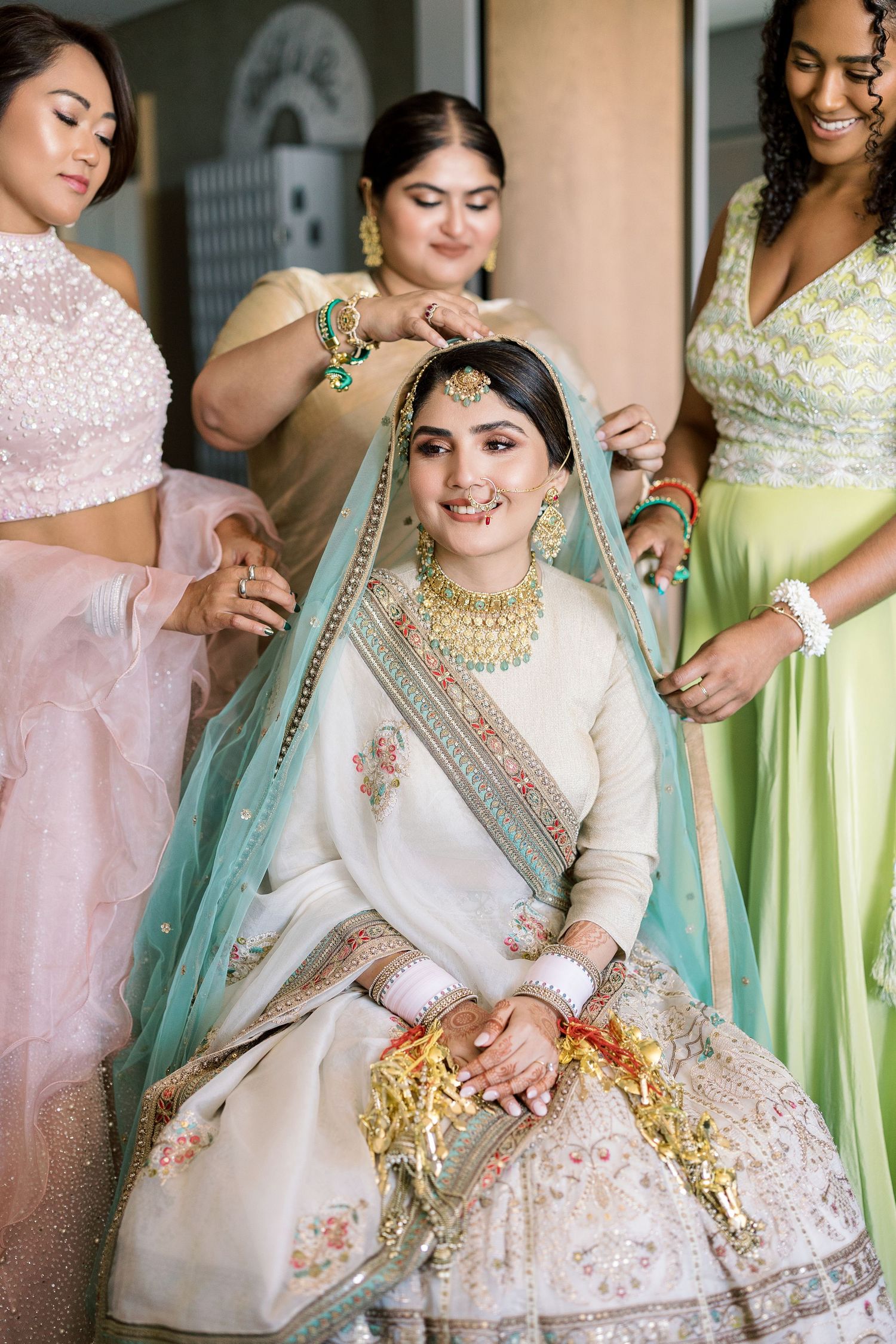 Traditional Indian bridal preparation scene with bridesmaids in pastel outfits helping with final touches.