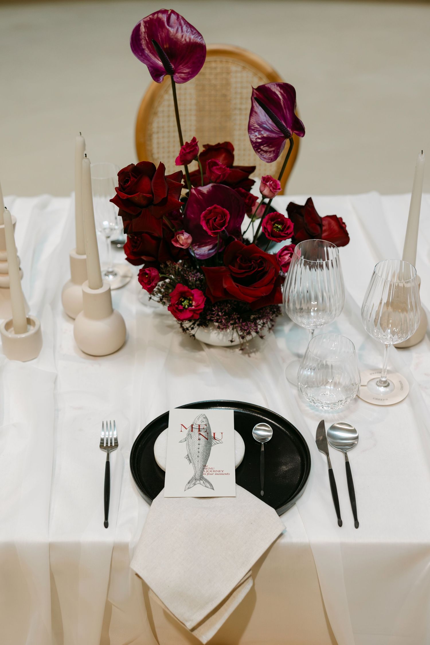 Elegant wedding table setting with red roses, anthurium blooms, black plates, white linens, and tapered candles.