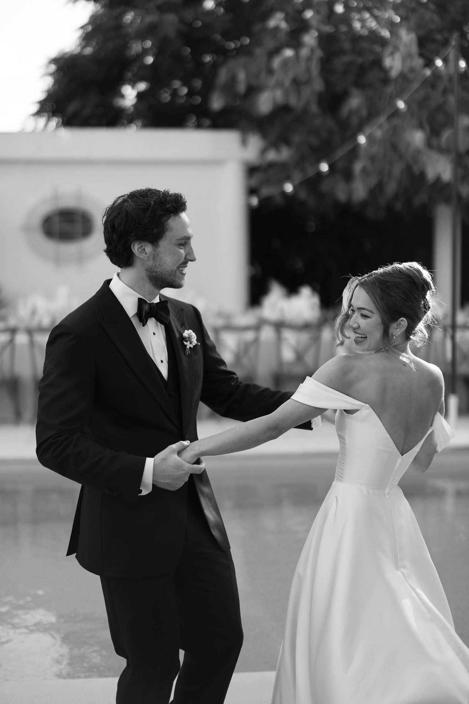Black and white photo of a smiling bride and groom holding hands and dancing outdoors near a pool at their wedding.