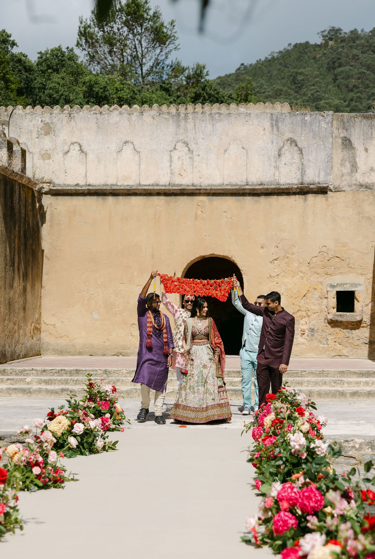 Pink roses bloom against a beige stone wall with arched doorways in a Mediterranean courtyard.