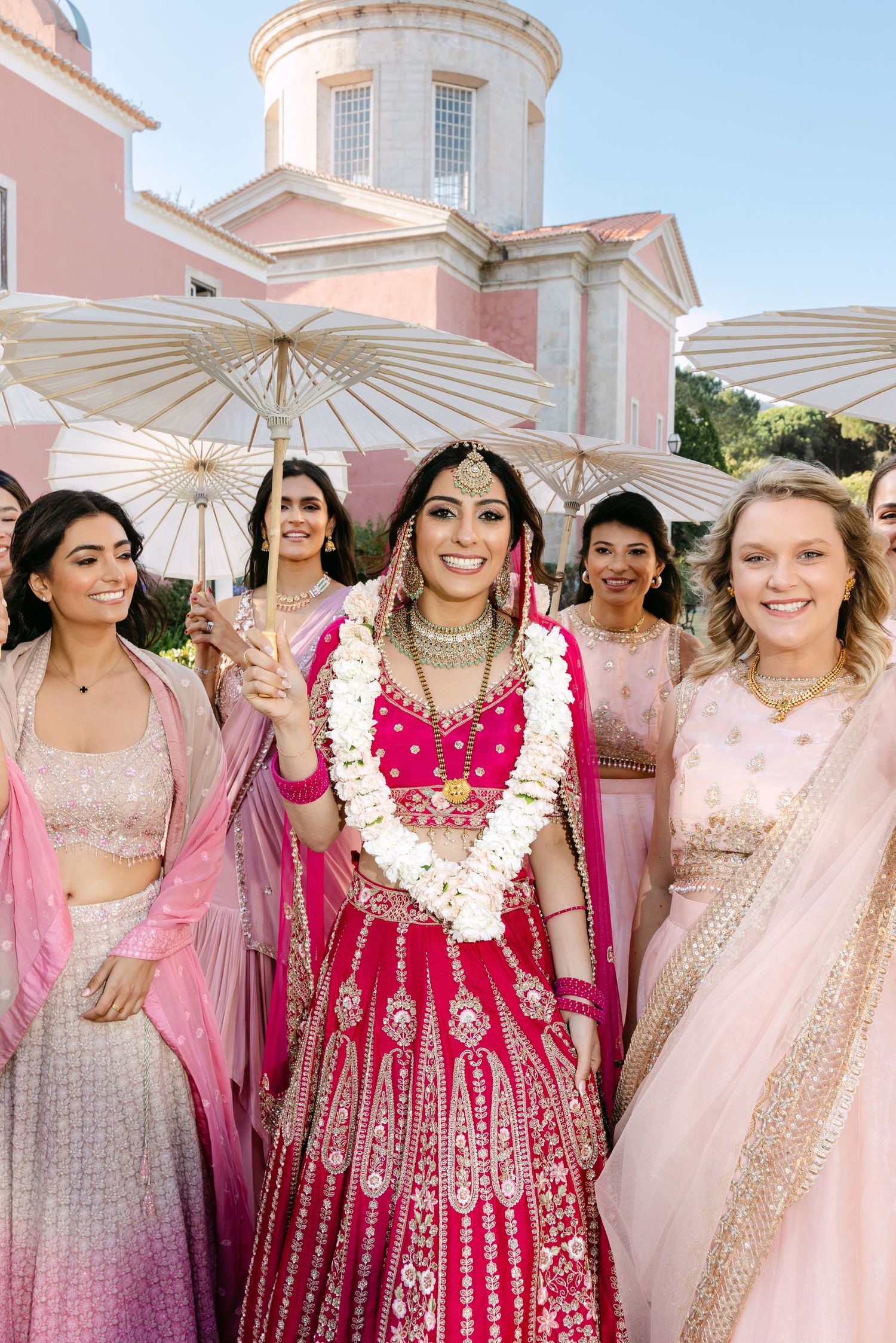 Bridesmaids in pink and cream outfits pose with parasols at an elegant wedding venue.