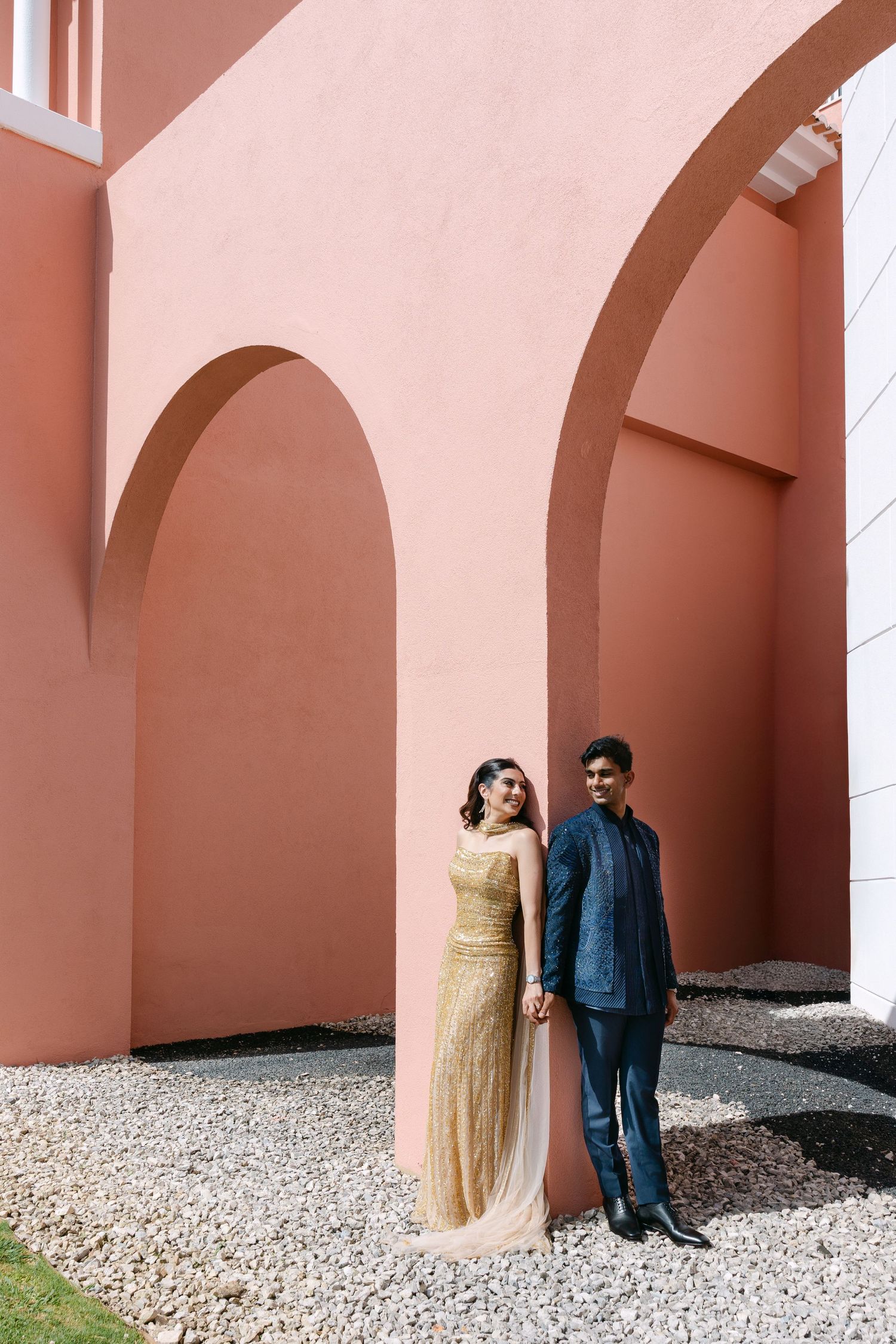 Couple in formal wear poses against coral-colored architectural arches.