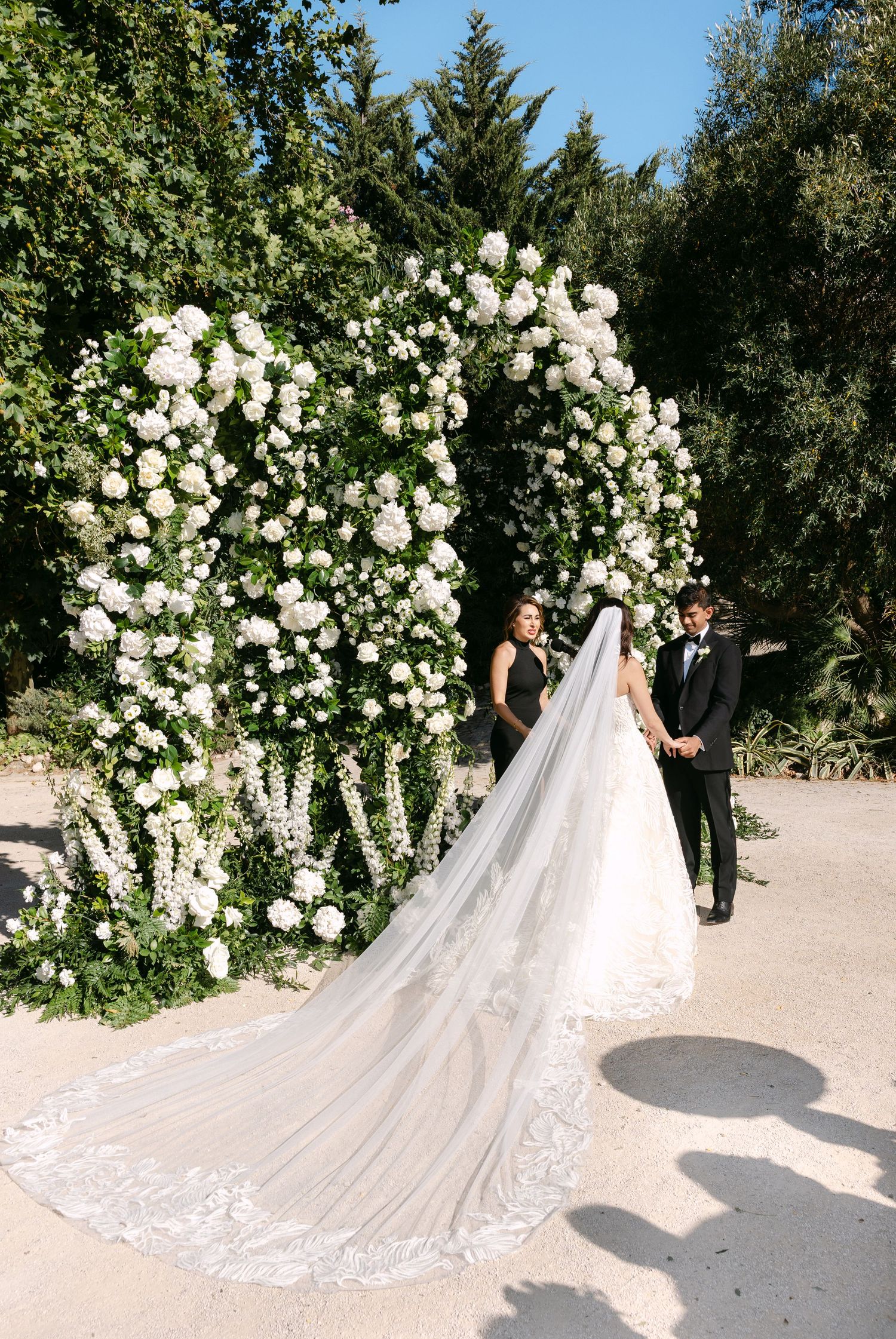 Wedding ceremony setup with white floral arch and long flowing train against a garden backdrop.