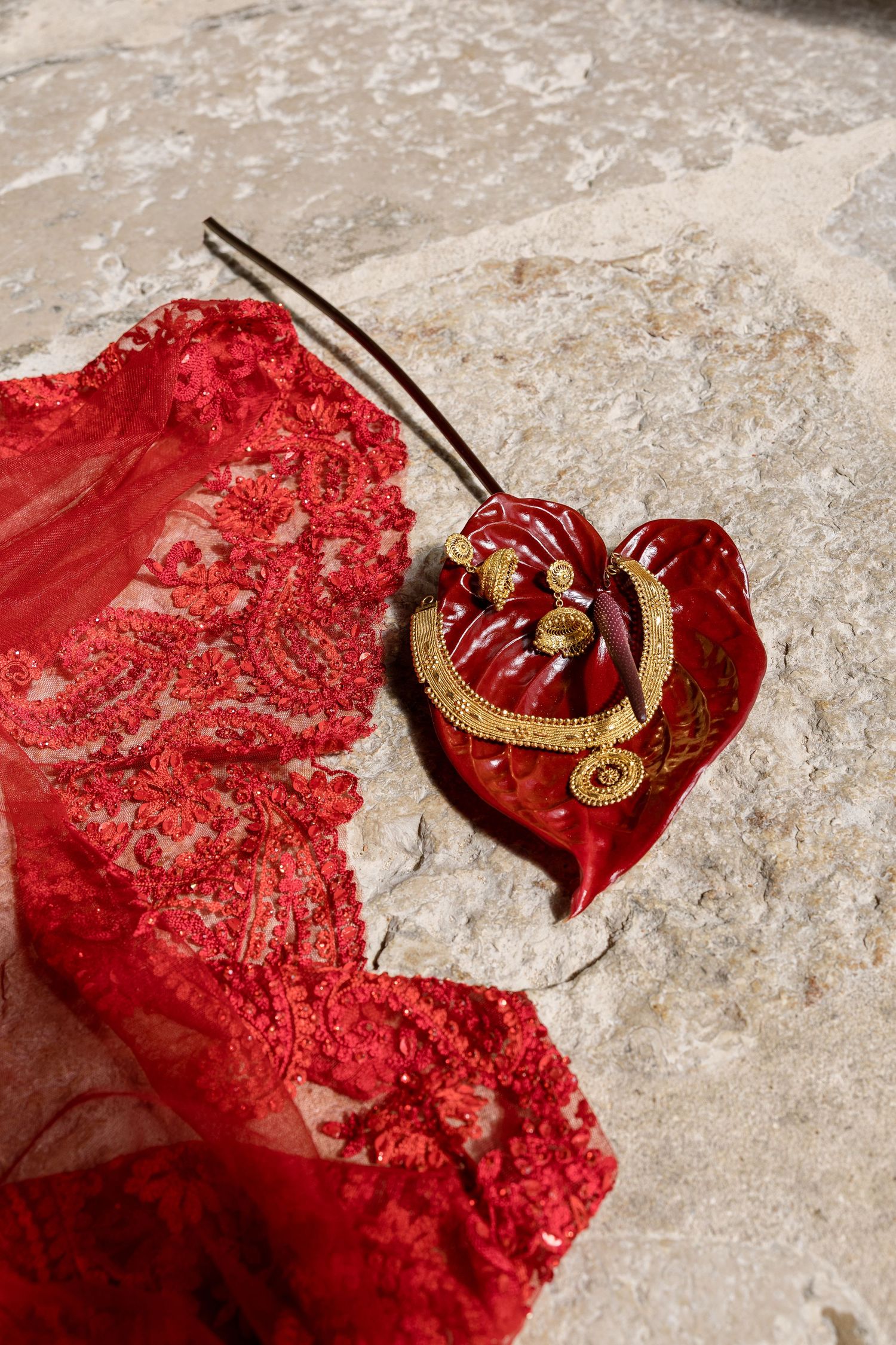Red lace fabric beside an anthurium flower with gold jewelry on stone surface.