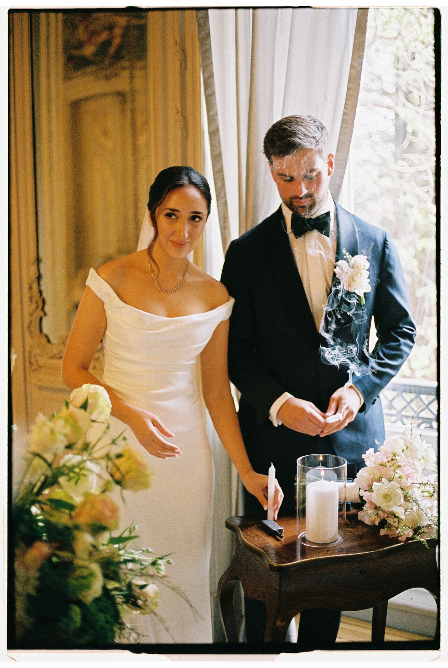 Bride and groom lighting a unity candle during their wedding ceremony, surrounded by white floral arrangements.