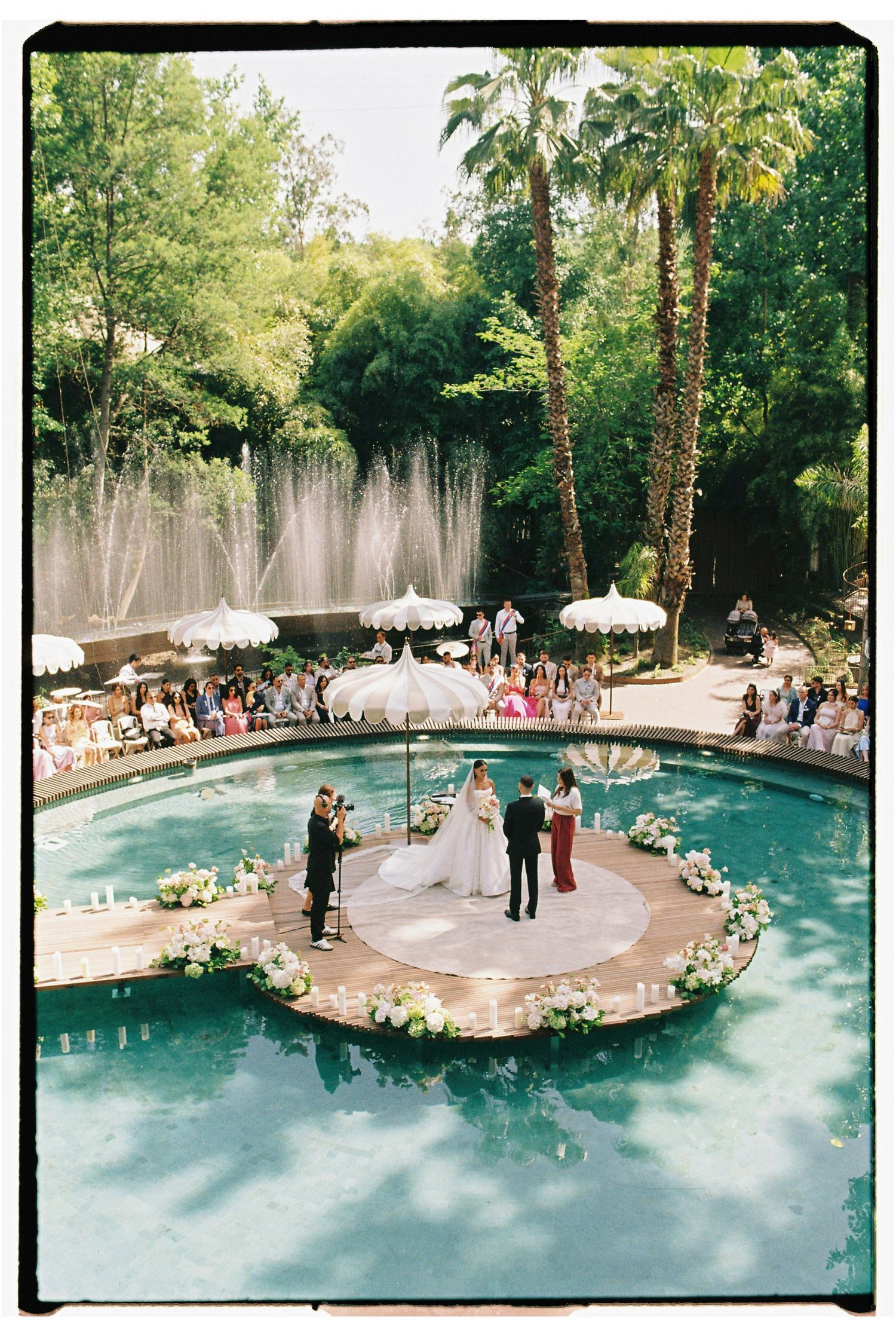 Outdoor wedding ceremony on a floating island platform in a pool, surrounded by lush greenery and fountains.