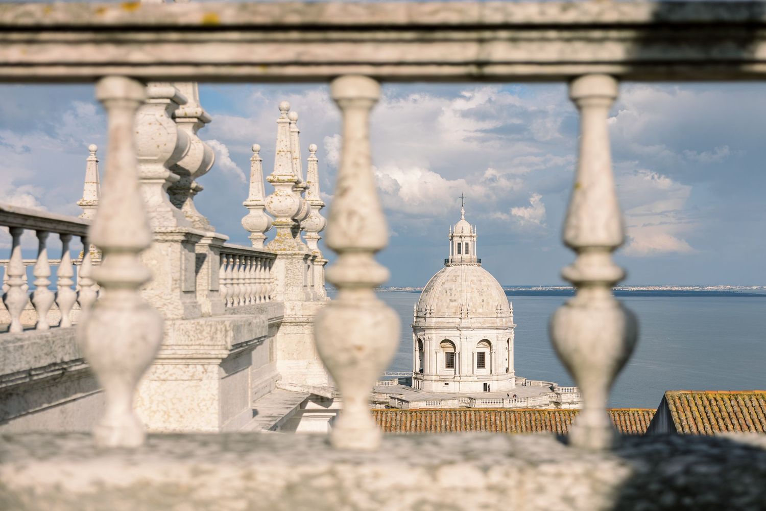 White stone balustrade with ornate columns framing a domed church and blue sky over Lisbon, Portugal.