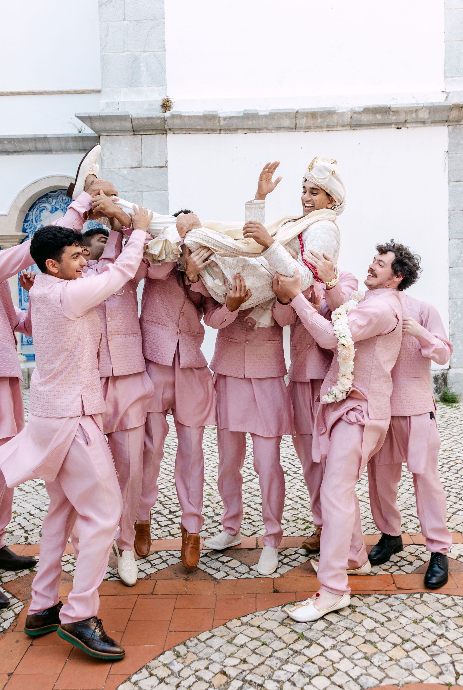 Groomsmen in matching pink suits playfully lift someone during a wedding celebration.
