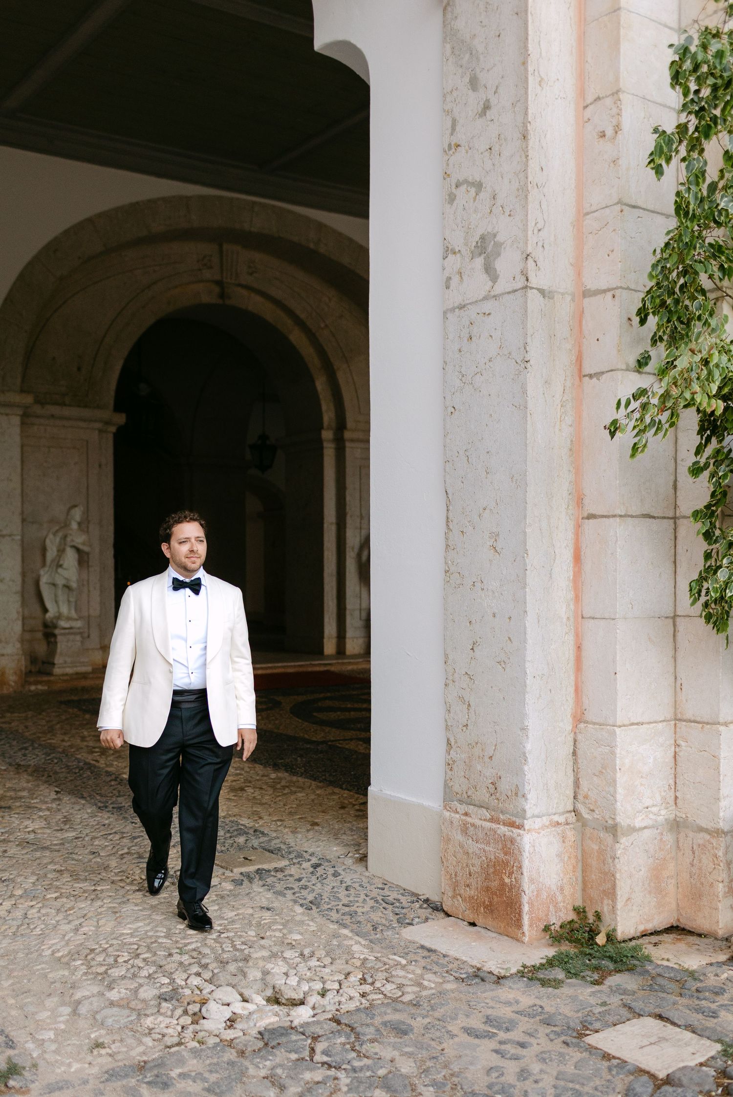 A person in white dinner jacket walks through stone archways in a historic setting.