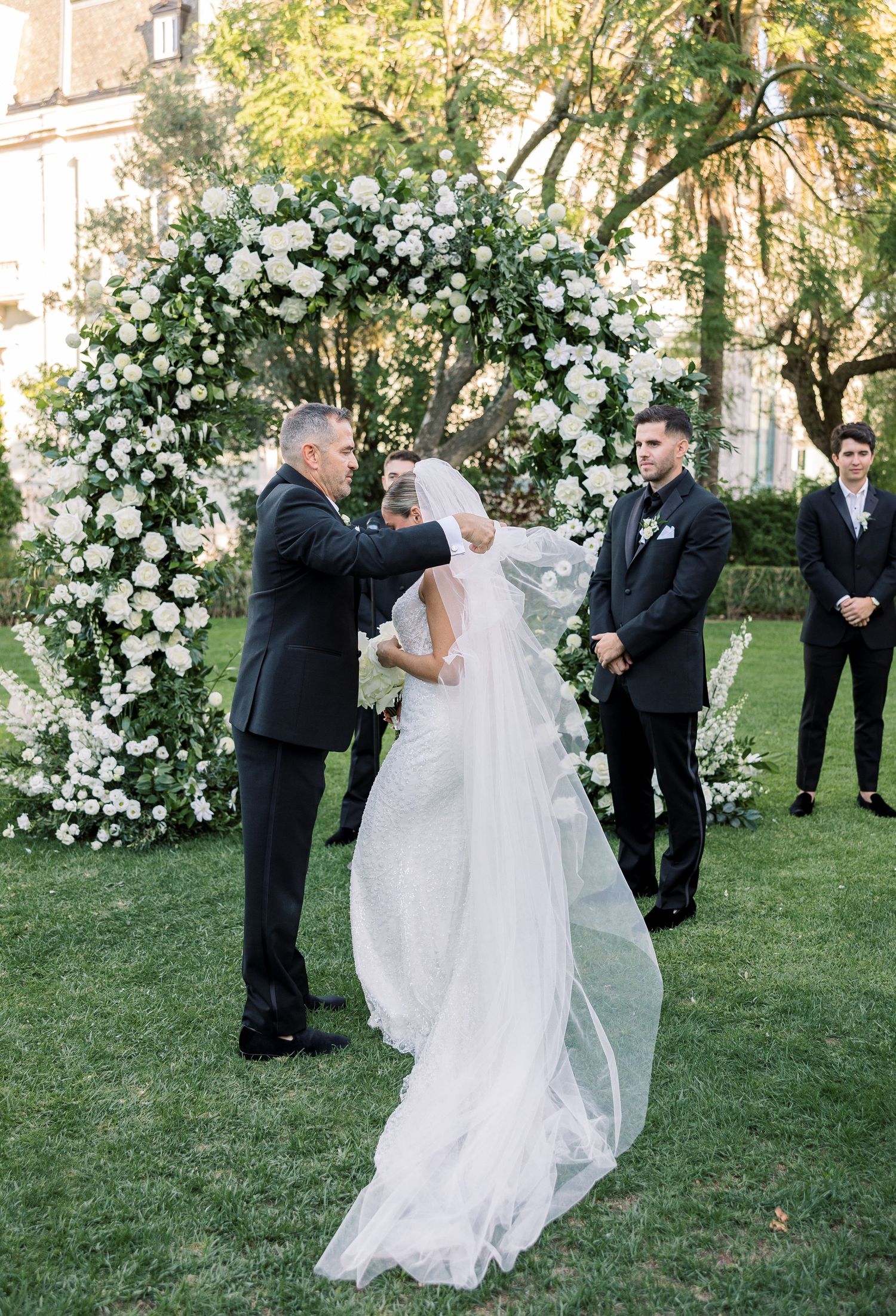 Wedding party poses on a lush lawn with elegant white floral arrangements and archway.