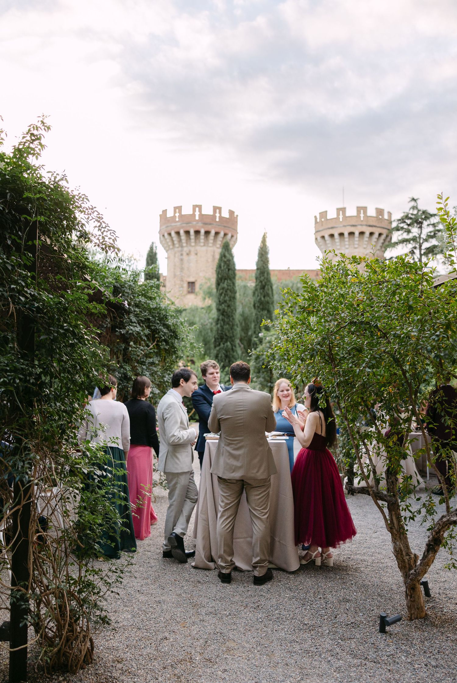 Wedding guests gather on a gravel path with castle towers visible in the background.