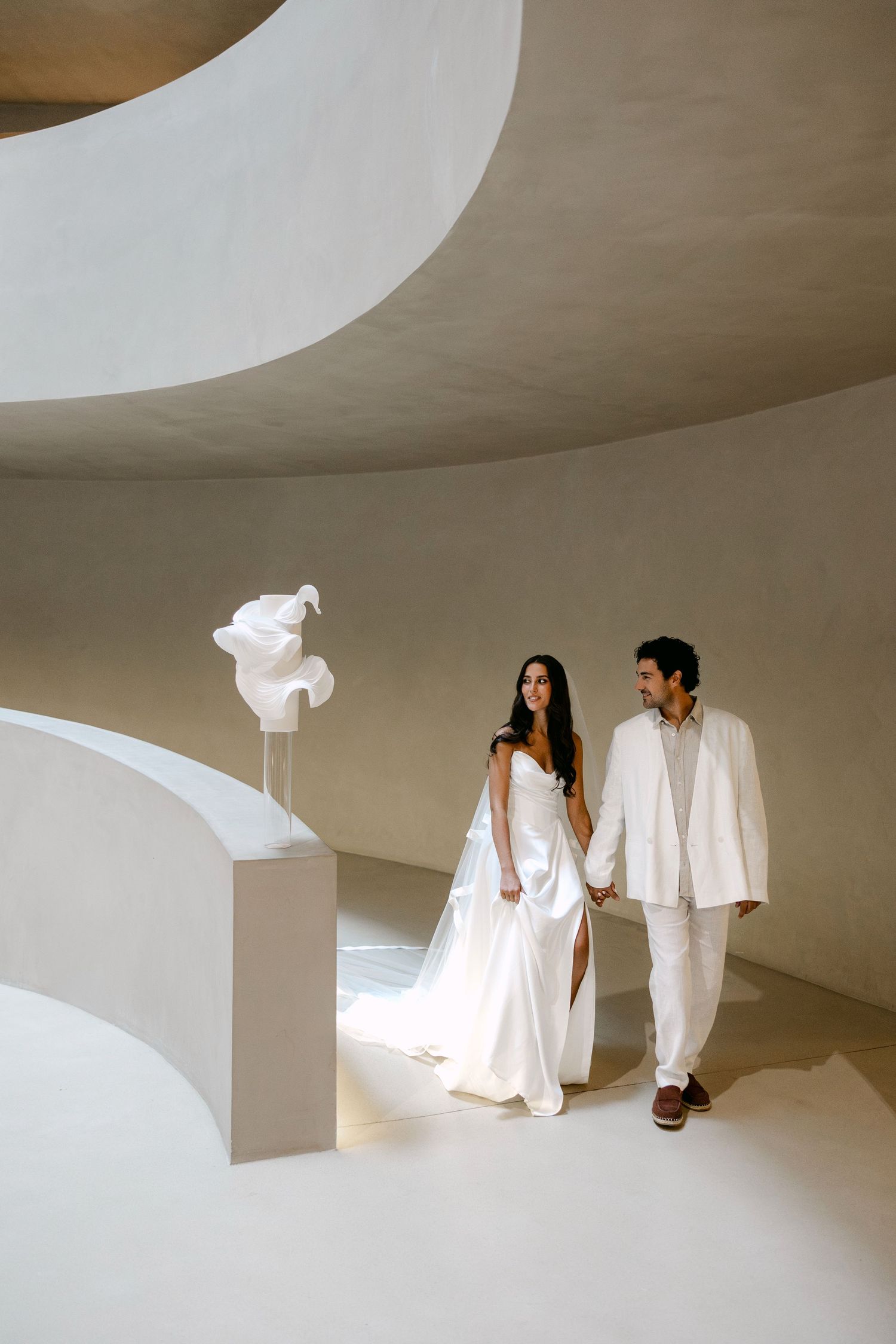 Bride and groom in white attire hold hands in a minimalist curved beige architectural space with sculptural decor.