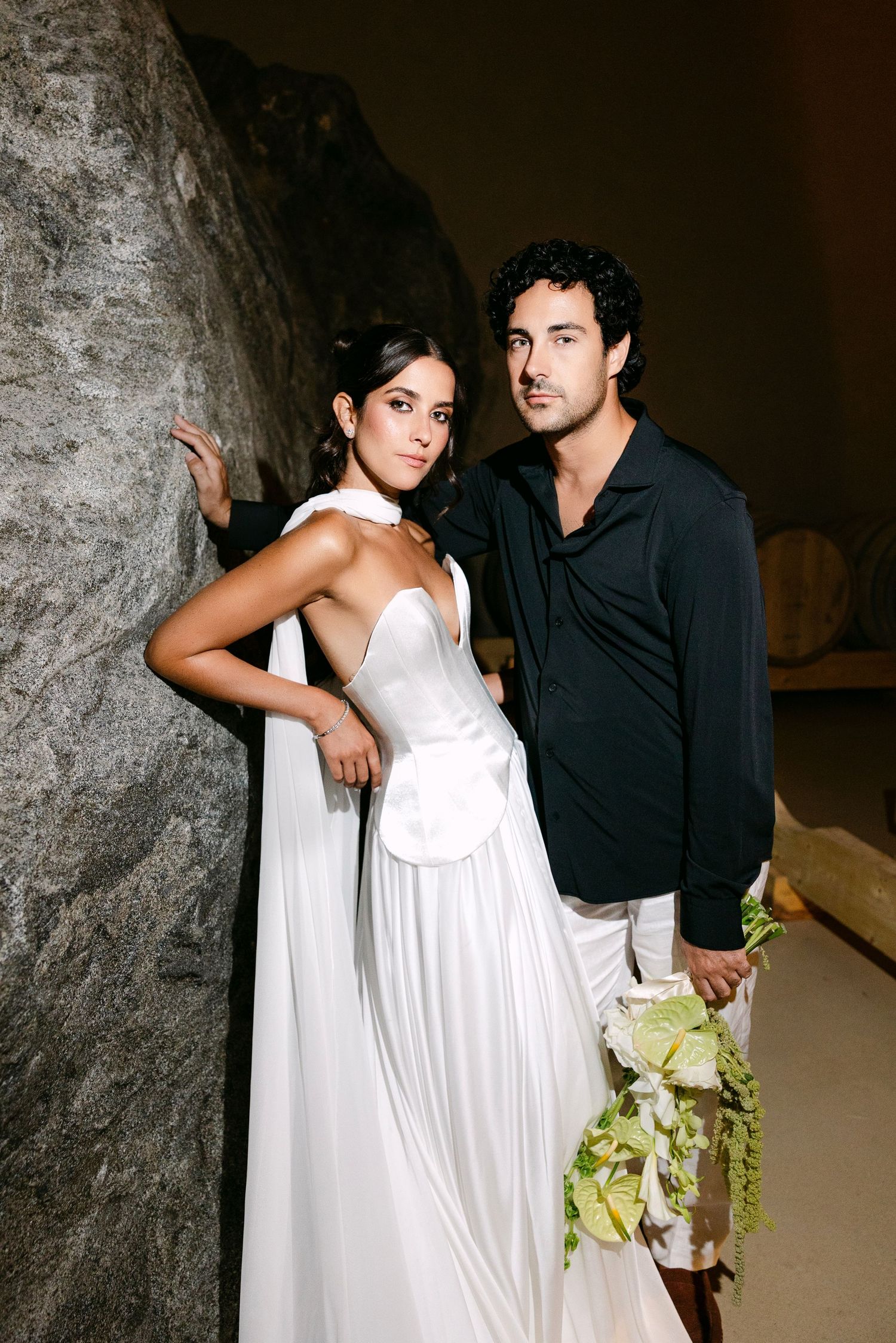 Bride in white gown and groom in black shirt pose against rocky wall holding a bouquet.