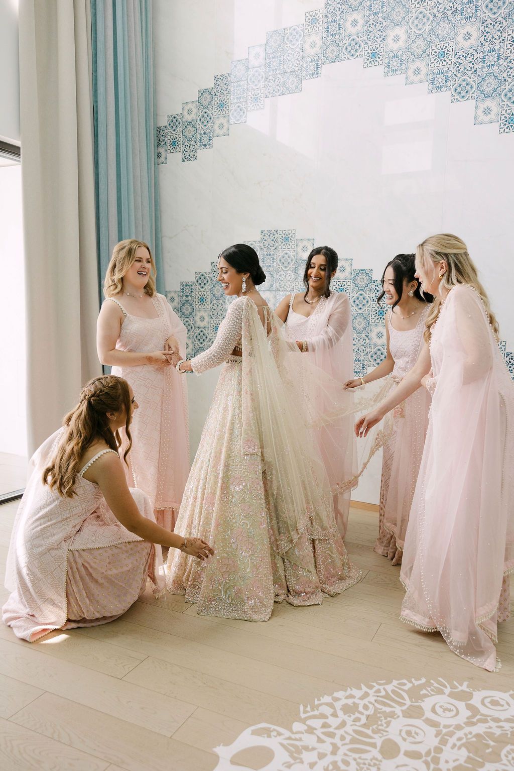 Bridesmaids in soft pink dresses help arrange a wedding gown in an elegant white room with blue wall details.