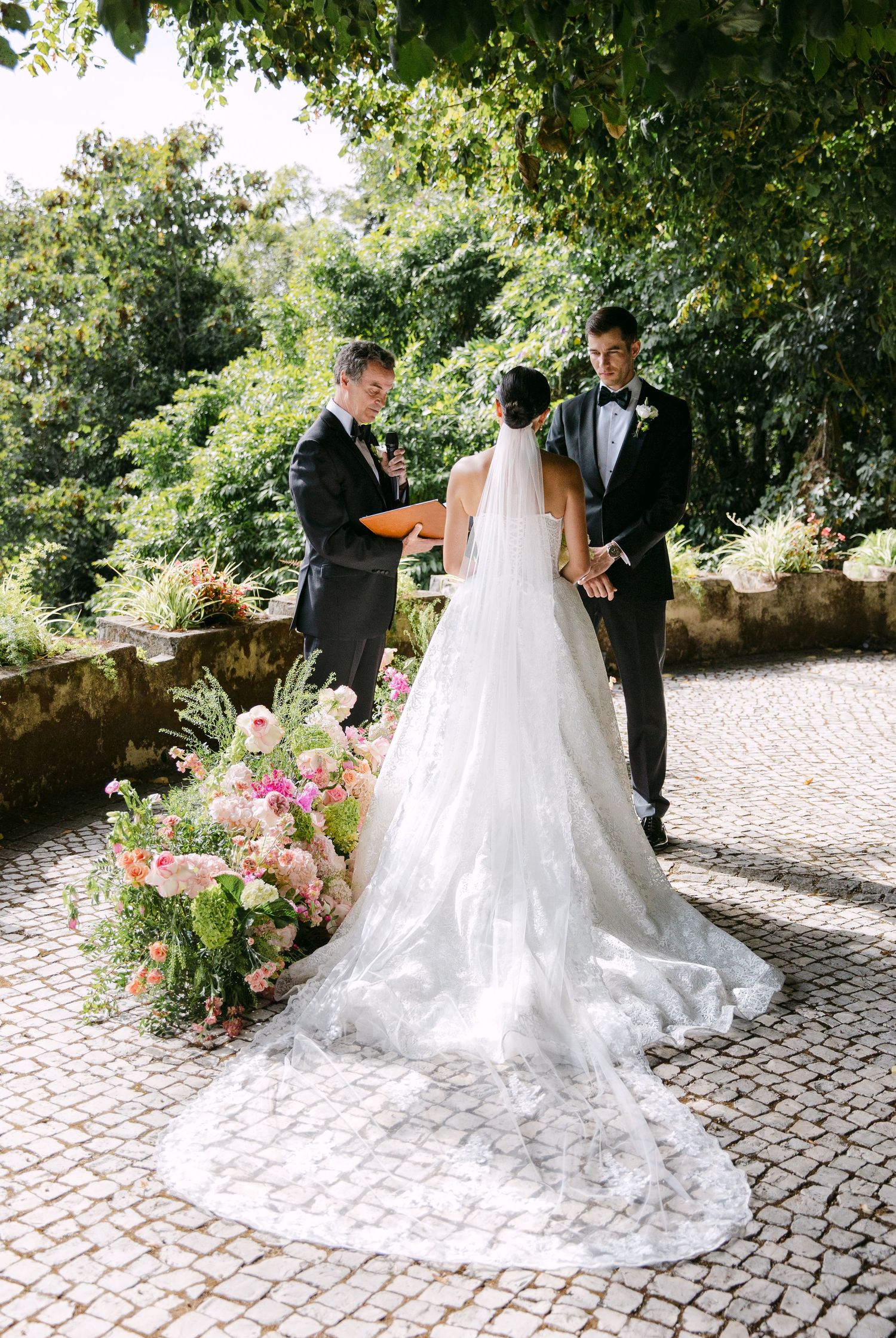 Wedding couple photographed from behind on cobblestone path with trailing lace dress and floral decor.