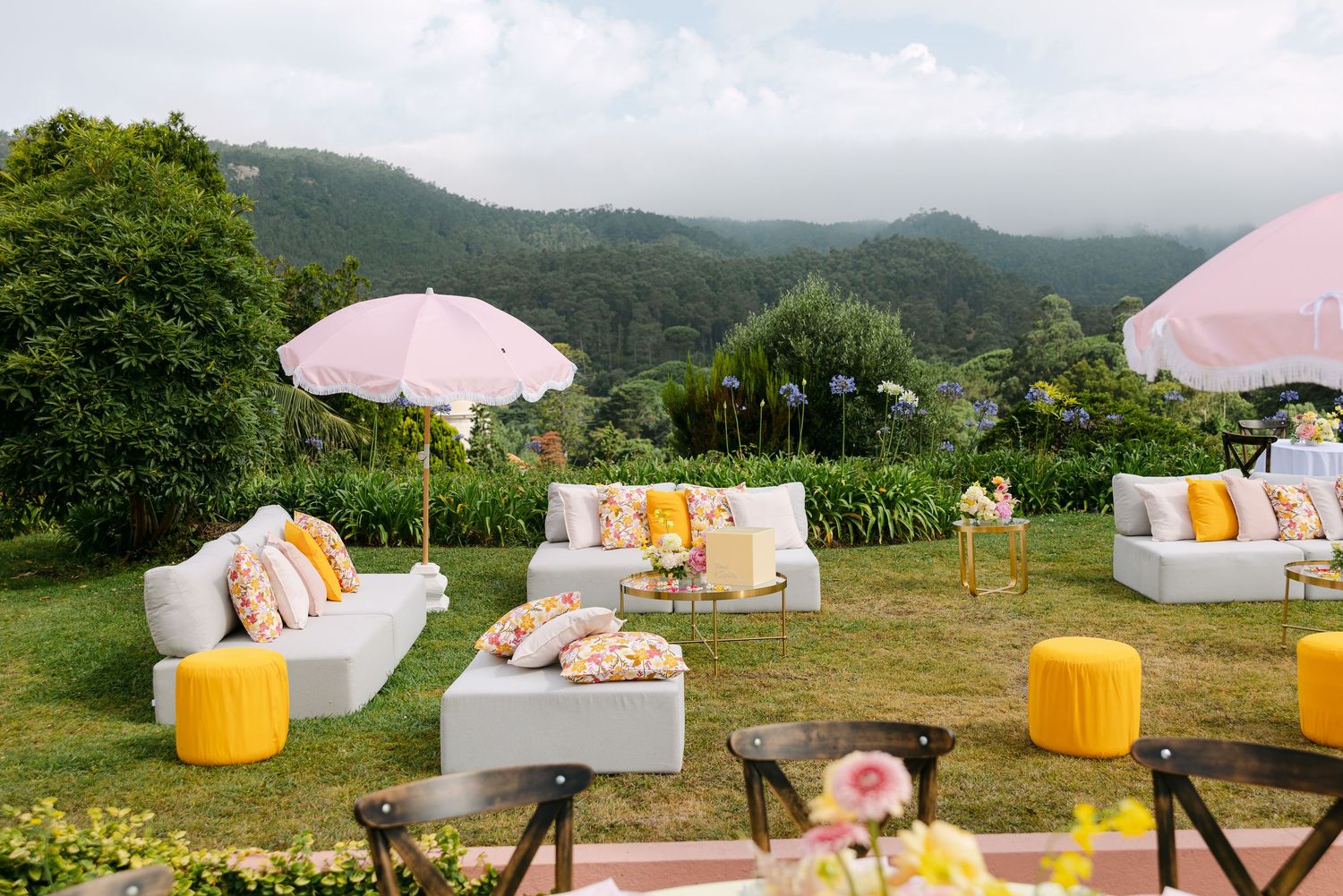 Outdoor wedding lounge area with white sofas, pink umbrellas and yellow accents against mountain backdrop.