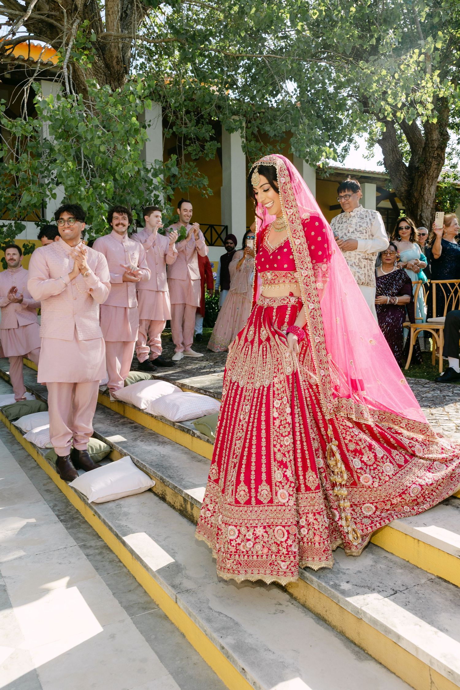 Wedding party in pink attire gathered outdoors with bride in traditional red Indian lehenga.