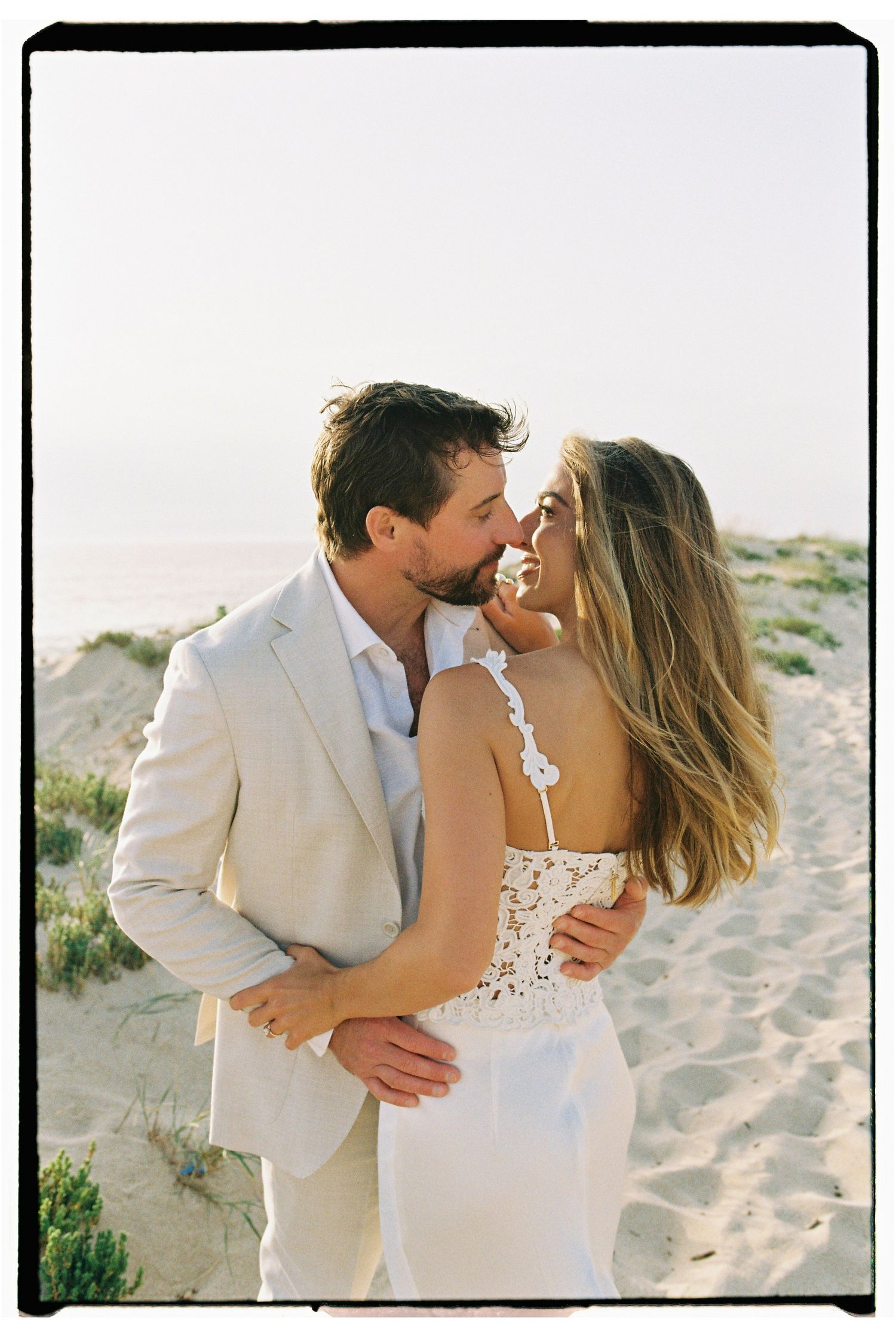 Romantic couple in wedding attire sharing intimate moment on sandy beach at sunset.