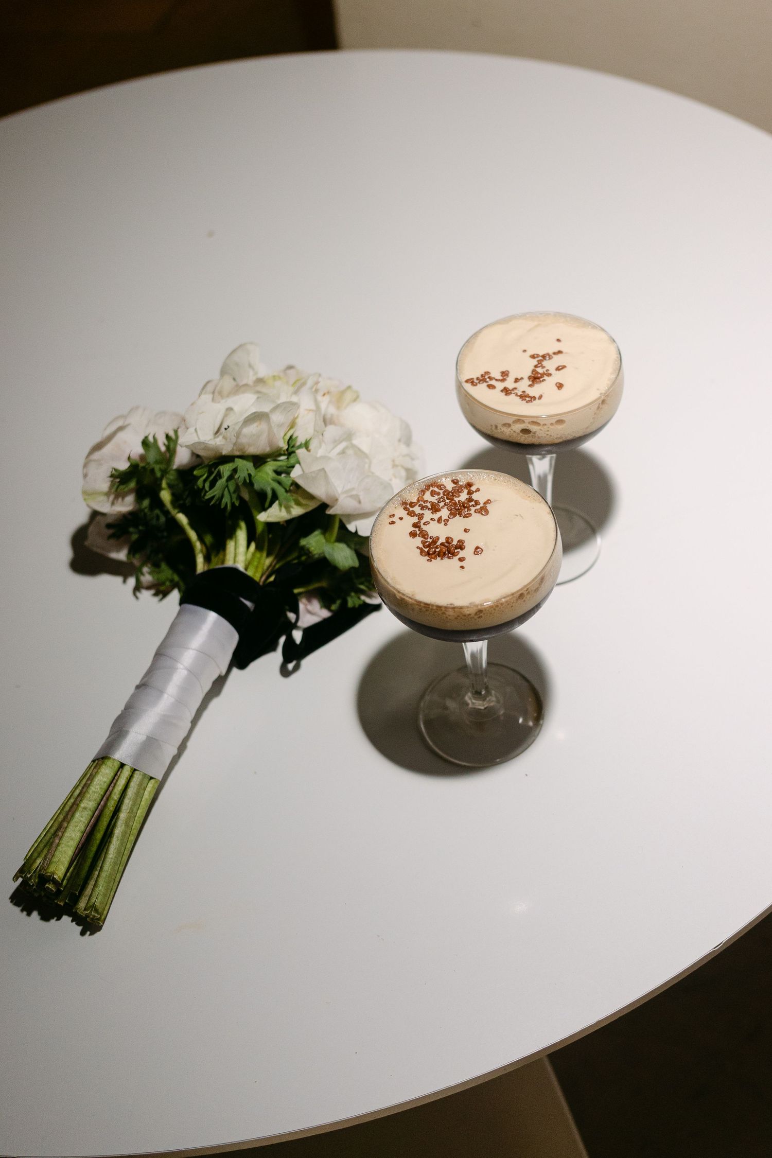 White floral bridal bouquet beside two espresso martini cocktails on white round table.