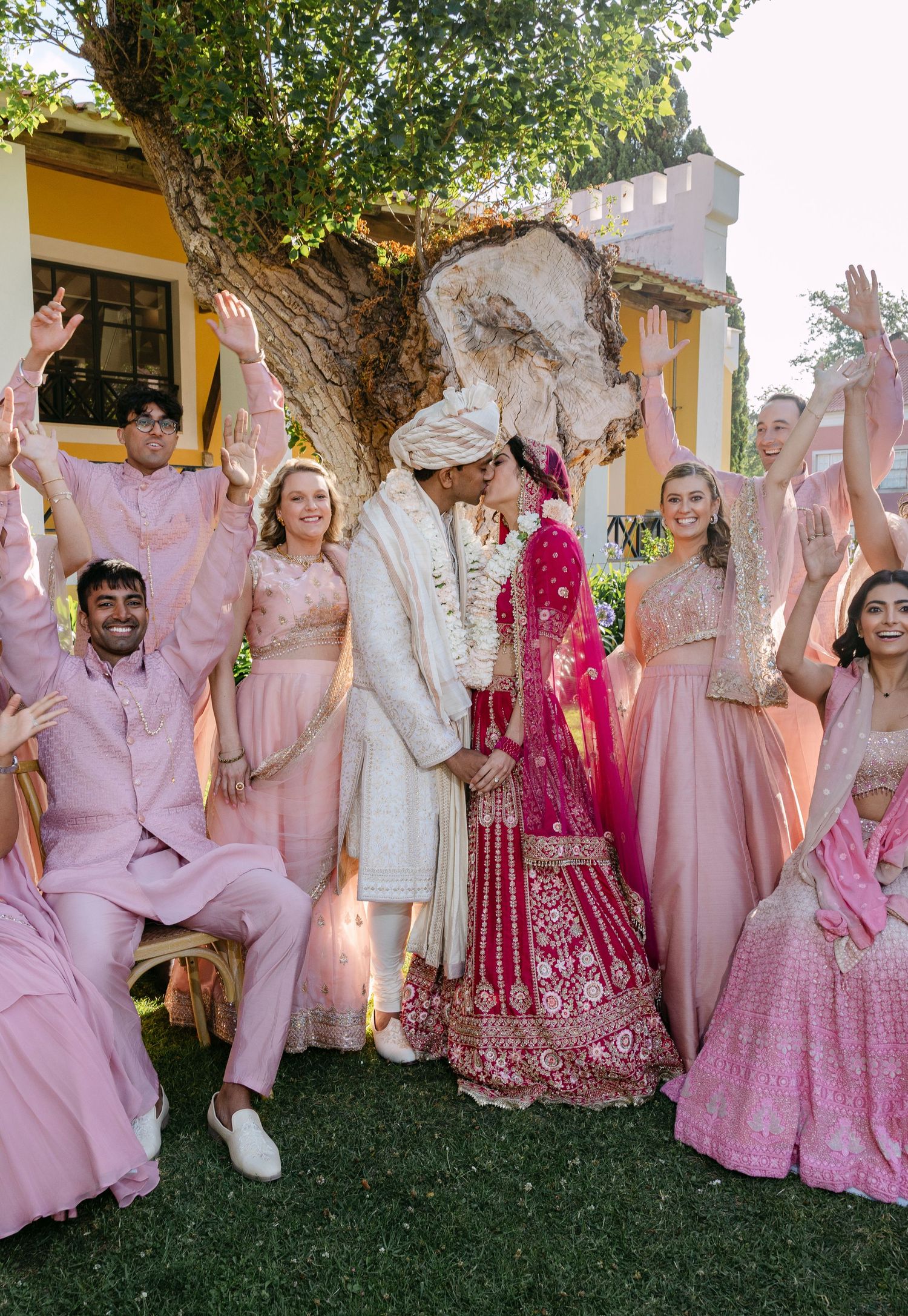 Wedding party in pink and traditional attire posing under a large tree during outdoor celebration.