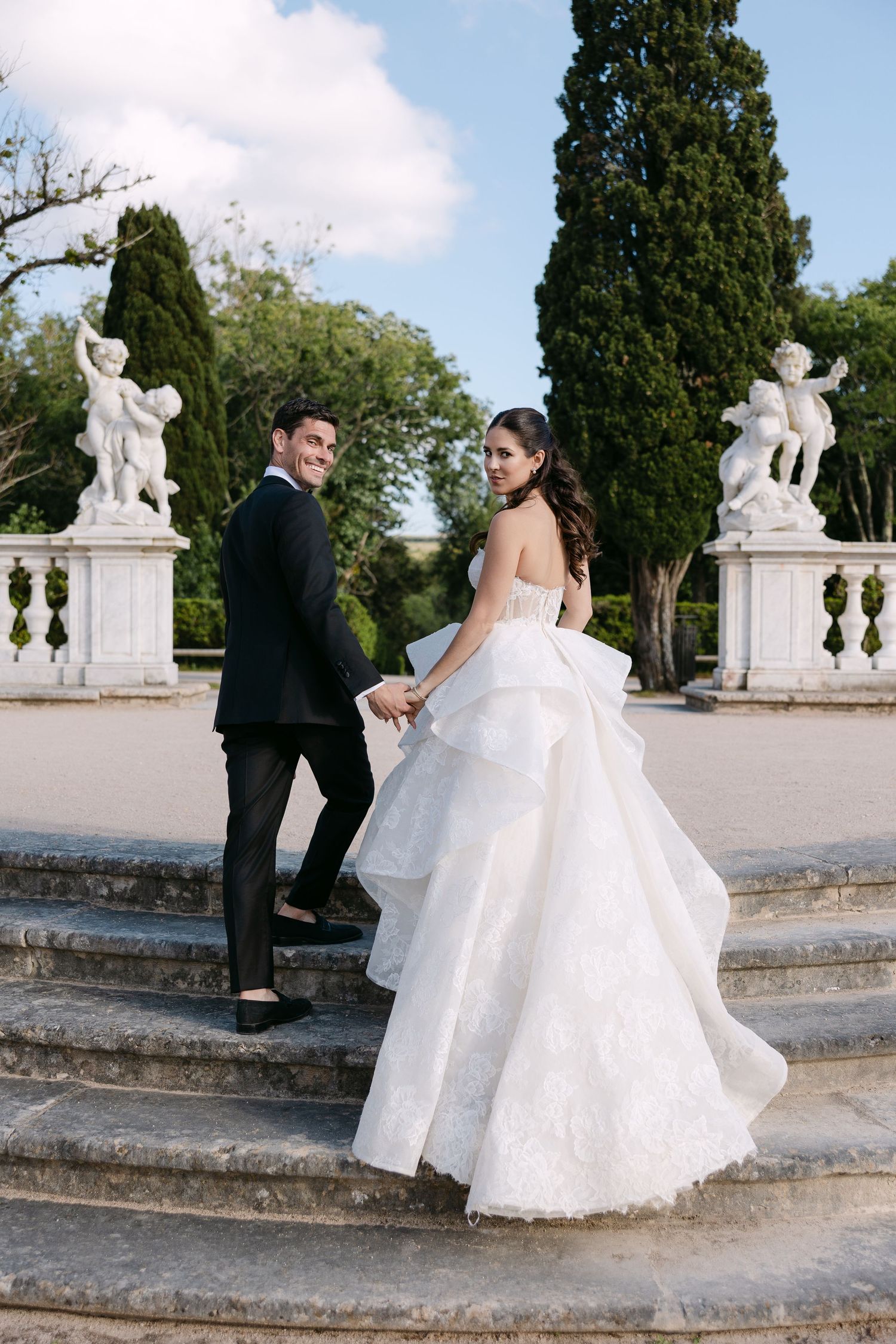 Romantic wedding couple poses on elegant stone steps surrounded by classical statues and cypress trees.