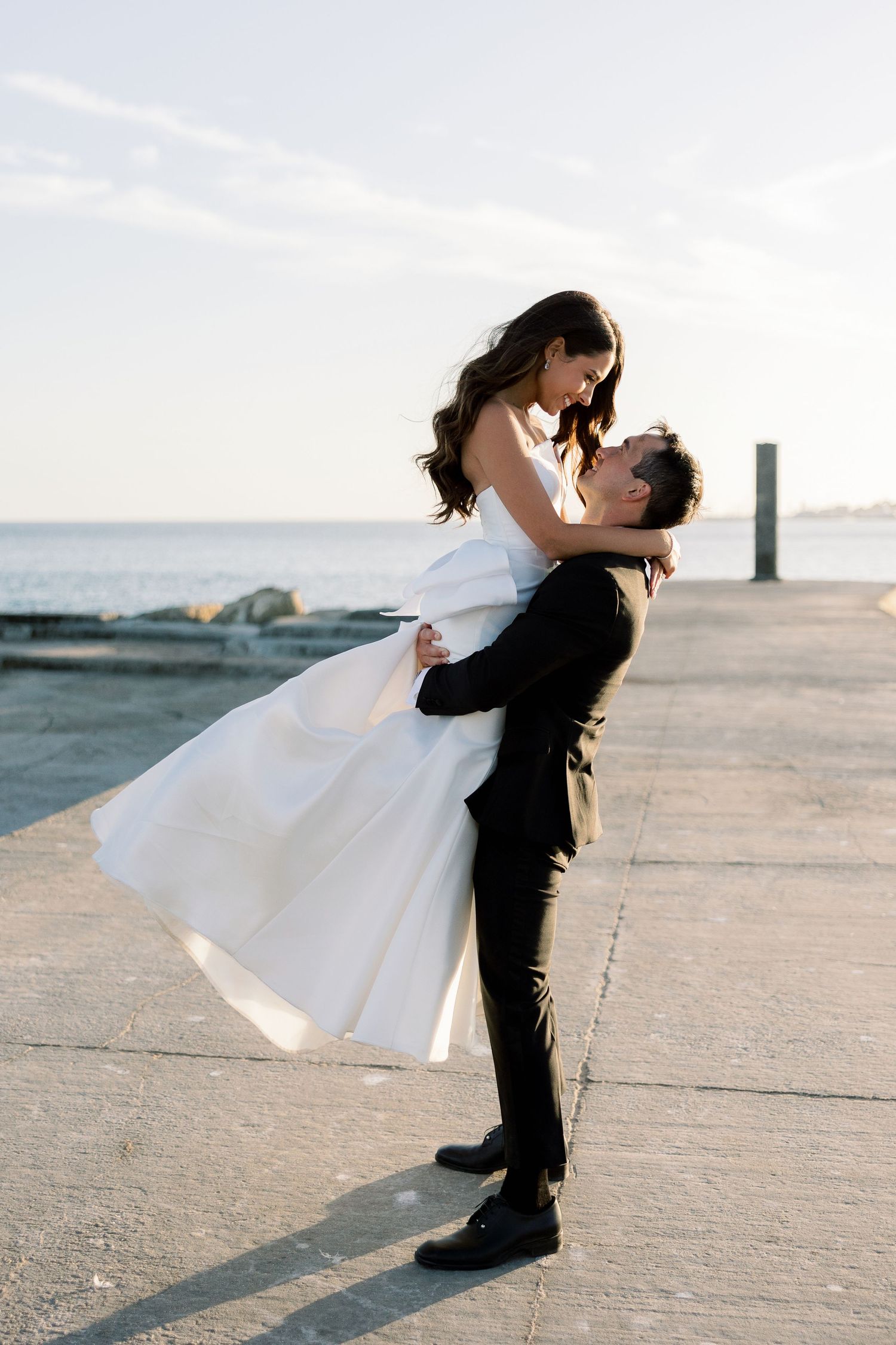 Couple in wedding attire embracing on a seaside pier during golden hour.