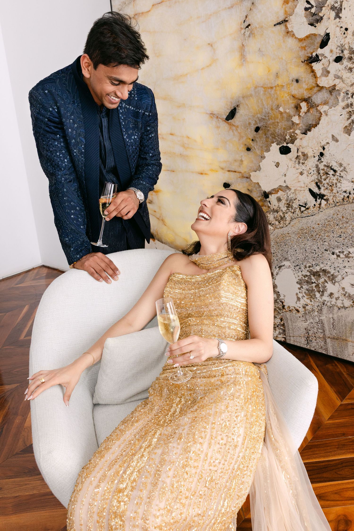 Couple shares champagne and laughter while relaxing on white couch, wearing formal navy suit and gold sequin dress.