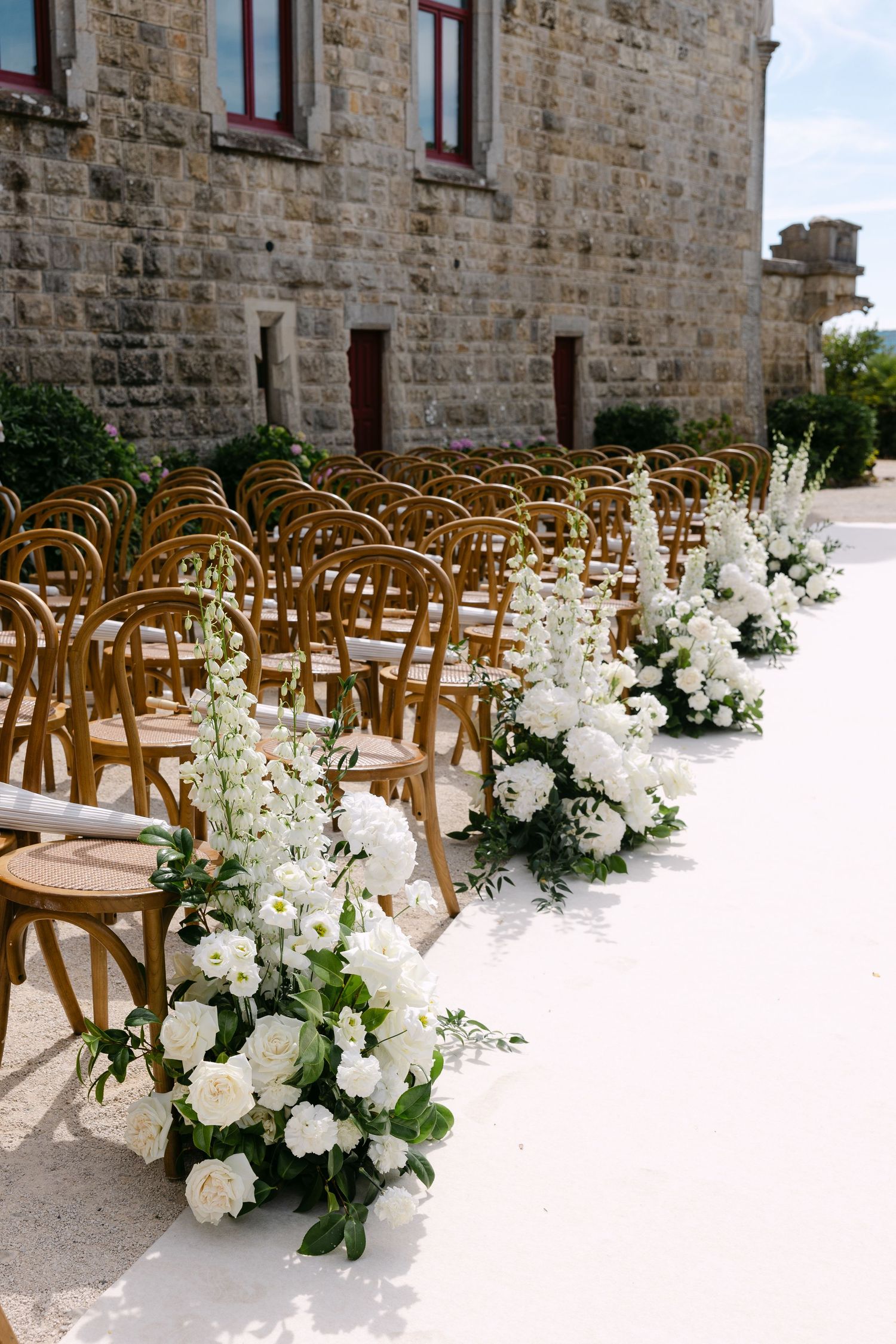 Wedding ceremony seating arrangement with wooden chairs and white floral decorations against stone wall.