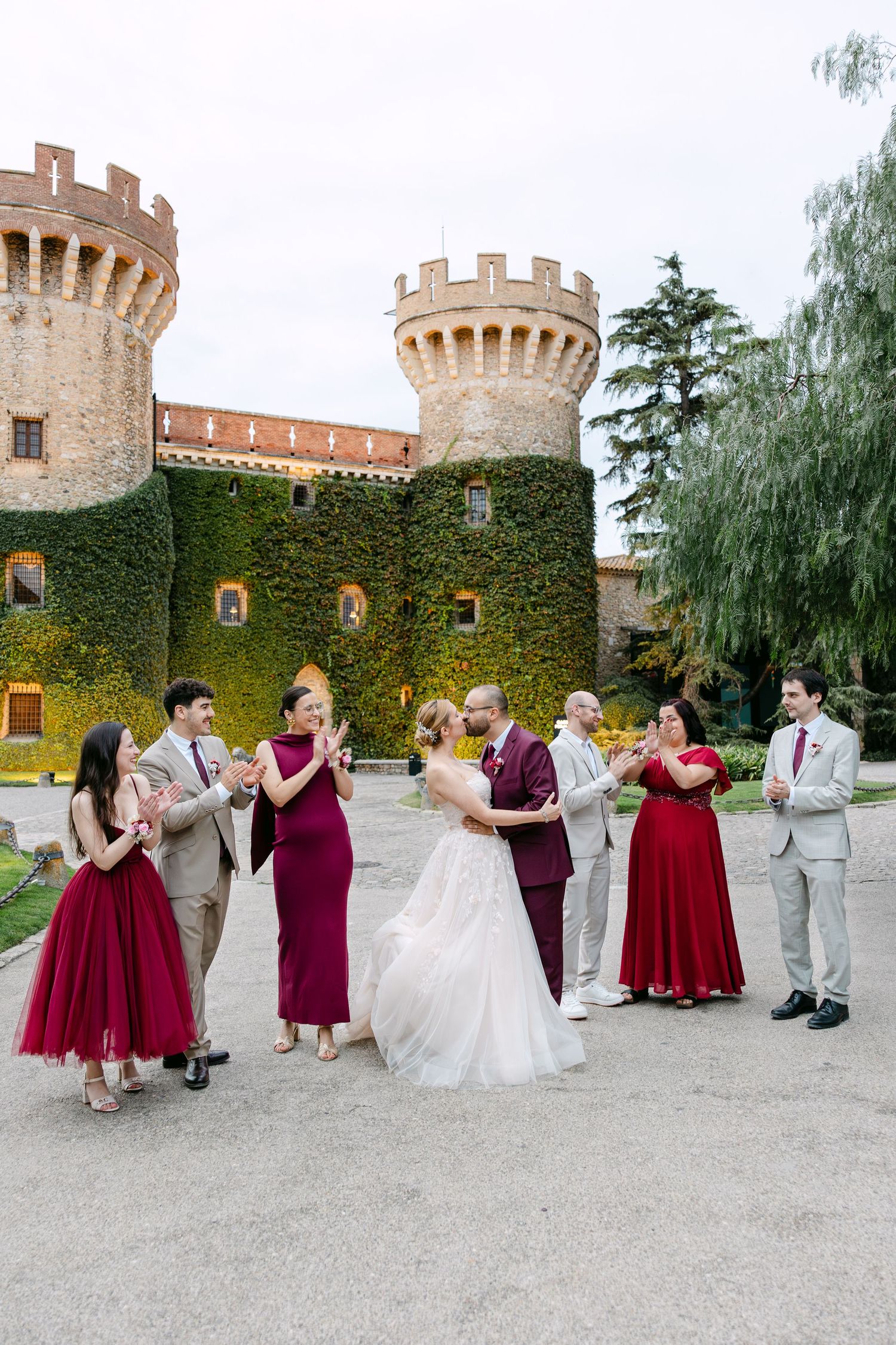 Wedding party in burgundy and gray attire poses in front of an ivy-covered castle wall with turrets.