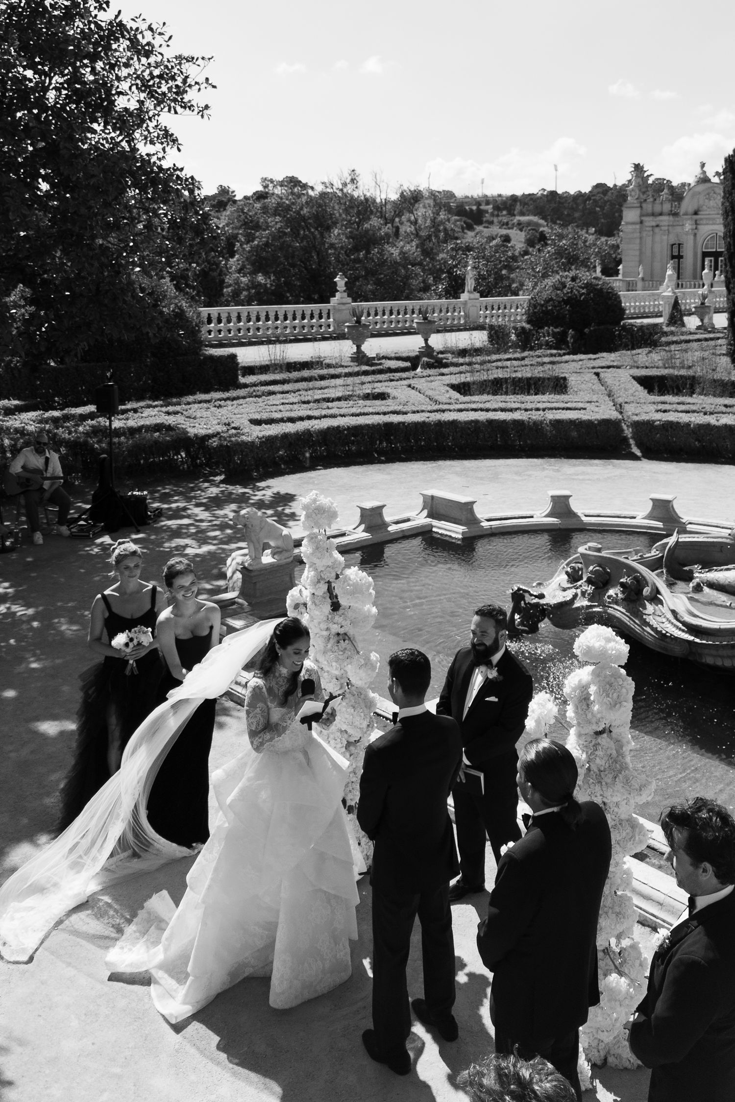 Bride and groom exchange vows at elegant outdoor wedding ceremony by ornate fountain.