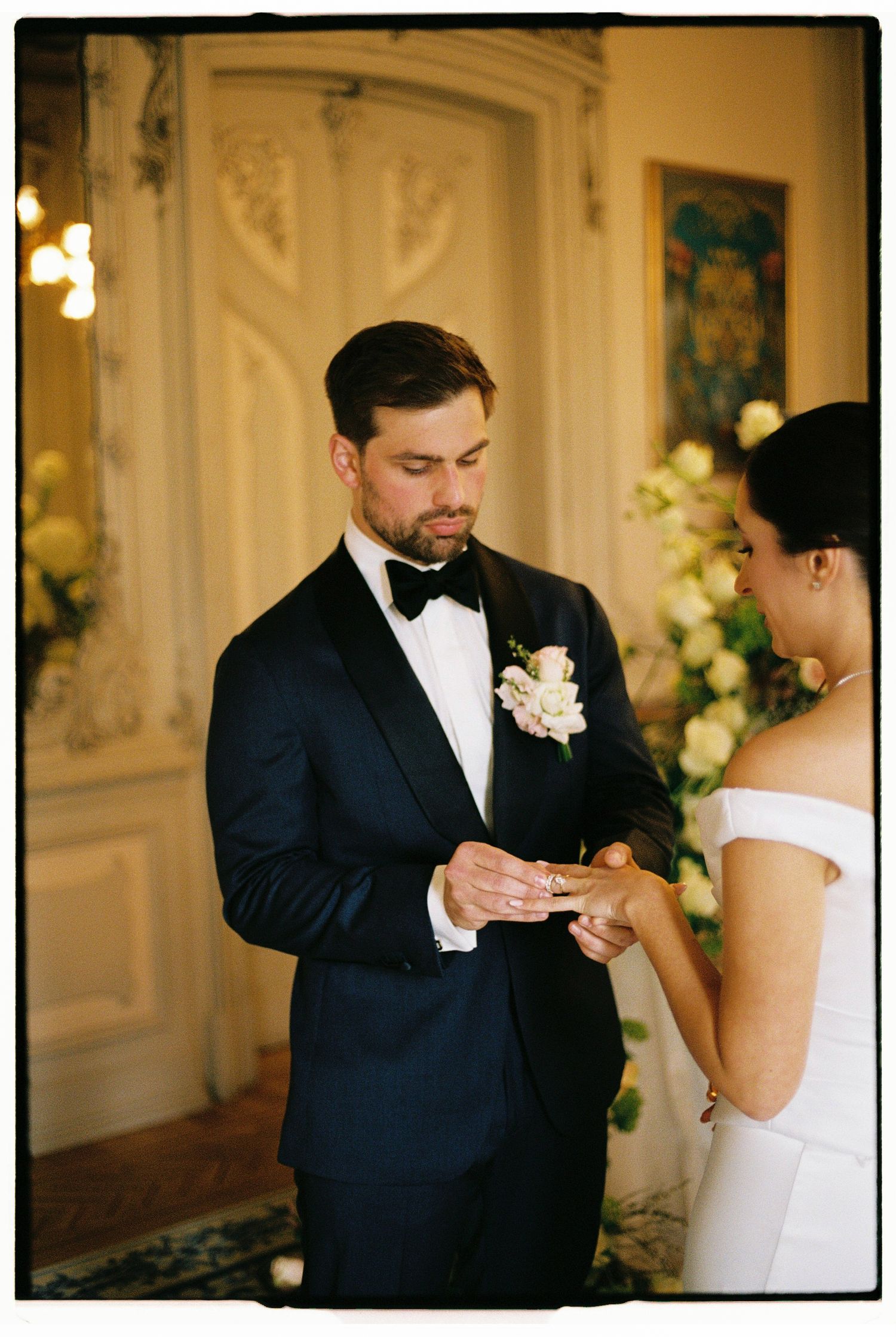 Groom in navy tuxedo placing wedding ring on bride's finger during elegant indoor ceremony with floral arrangements.