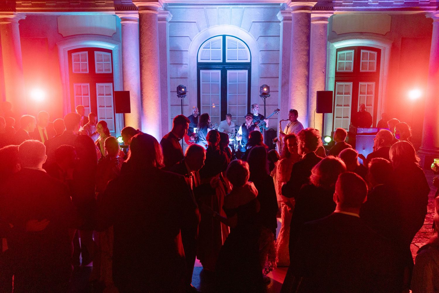 Wedding guests dance under colorful red and blue lights in grand classical venue.
