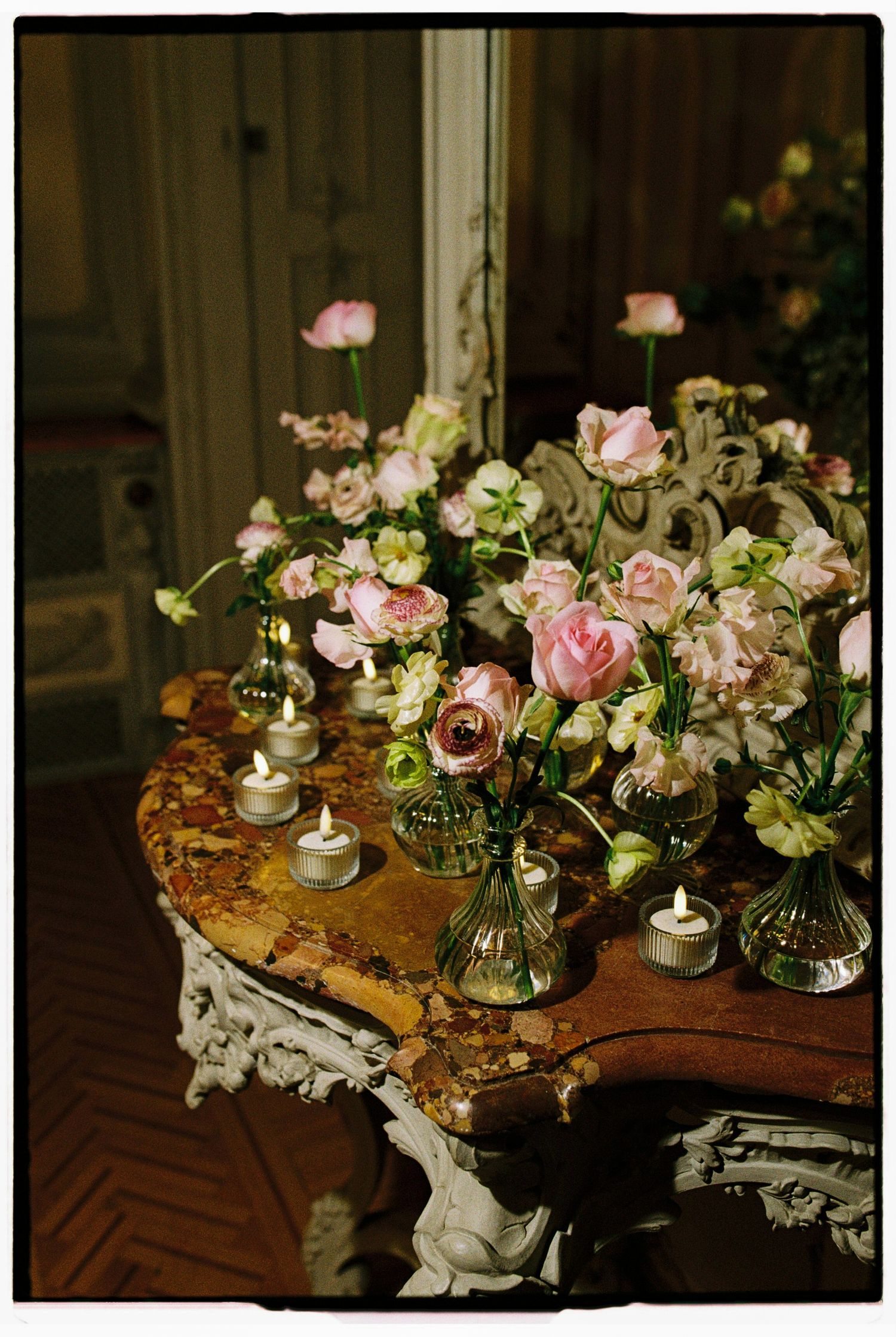 Elegant antique table with pink roses in glass vases and lit tea candles in a vintage interior setting.