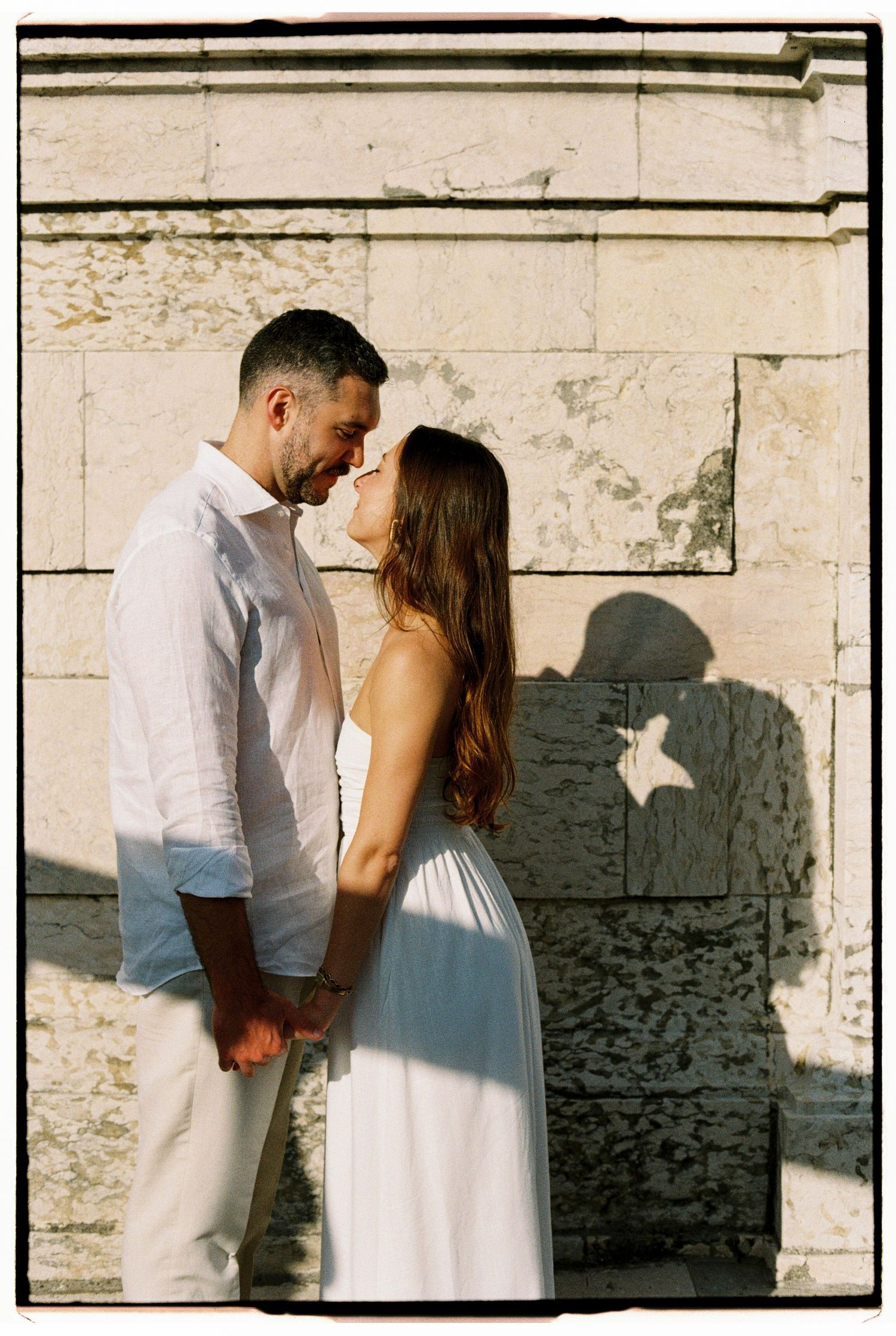 Romantic couple in white outfits holding hands and leaning in for a kiss against a sunlit stone wall.