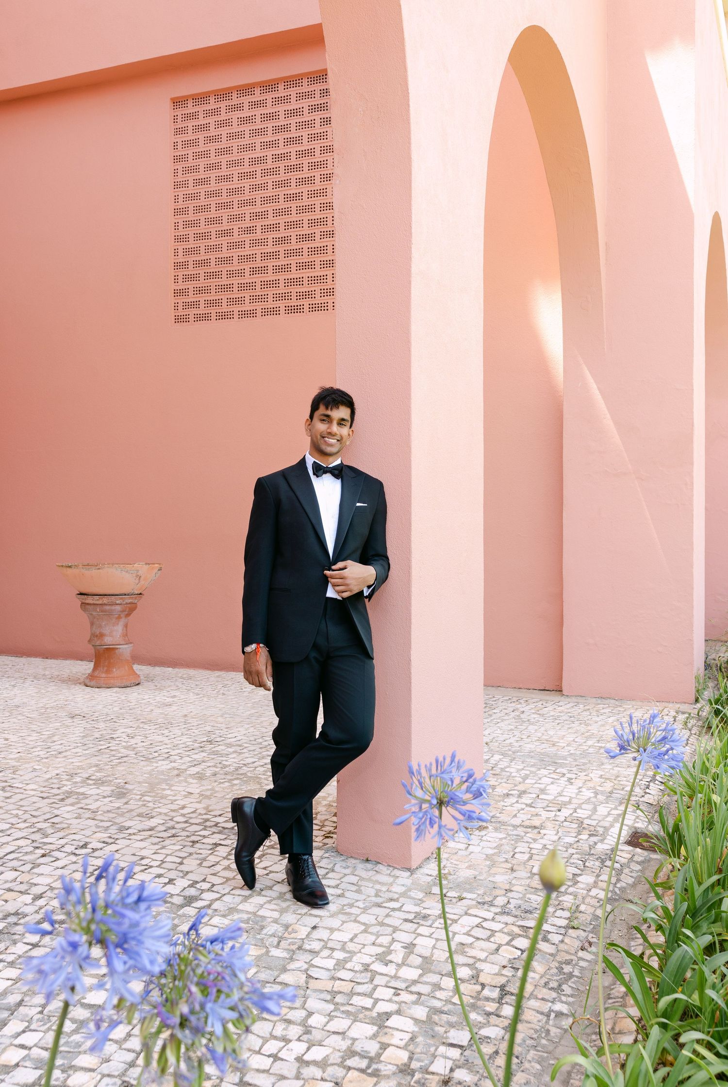 Groom in black tuxedo posing against coral pink wall with blue flowers in foreground.
