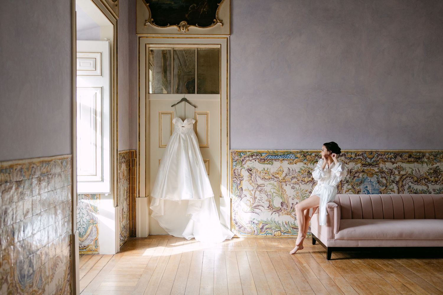 Sunlit room with vintage furniture and wedding dress displayed near large windows.