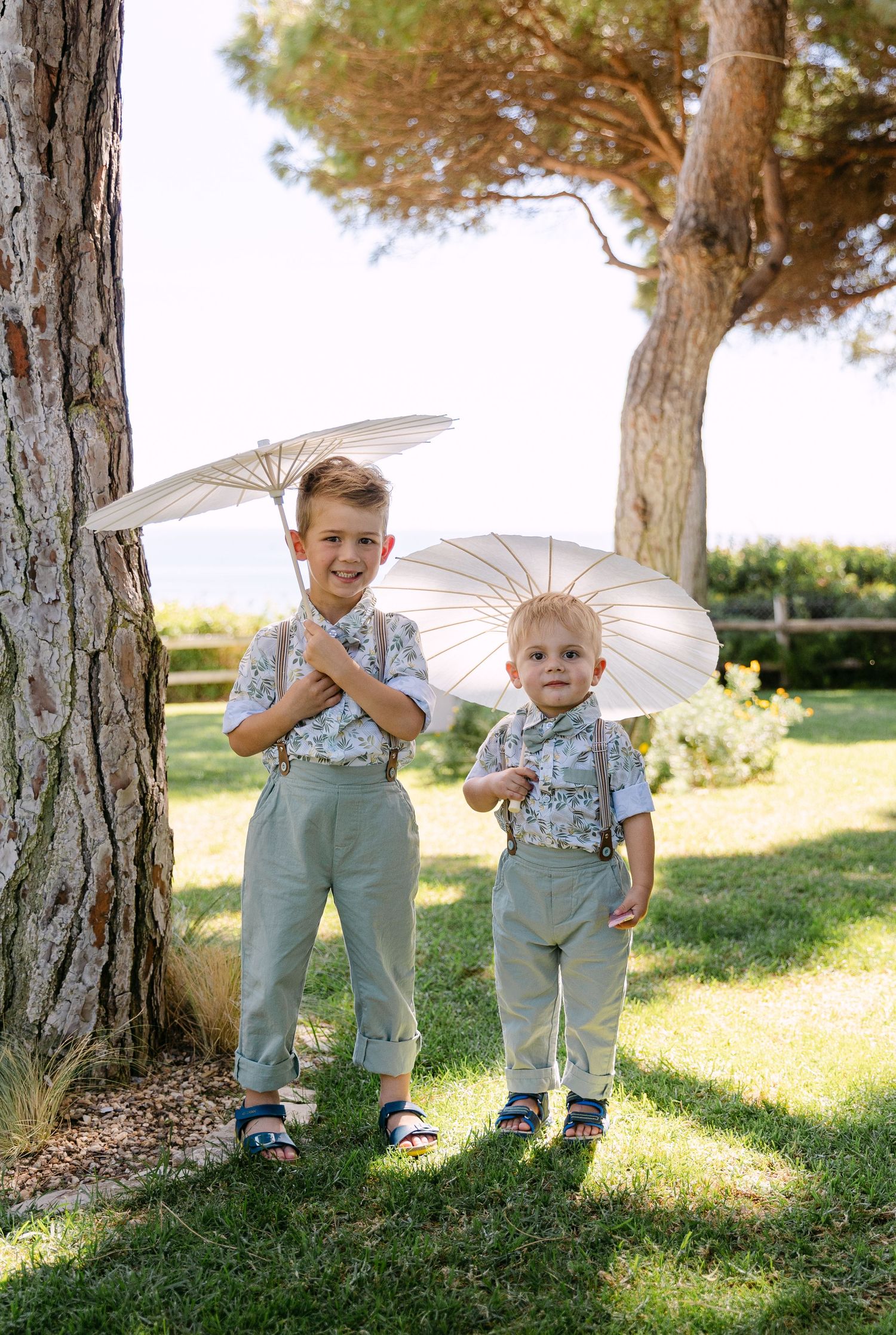Young children in overalls holding white parasols pose under pine trees in a sunny garden.