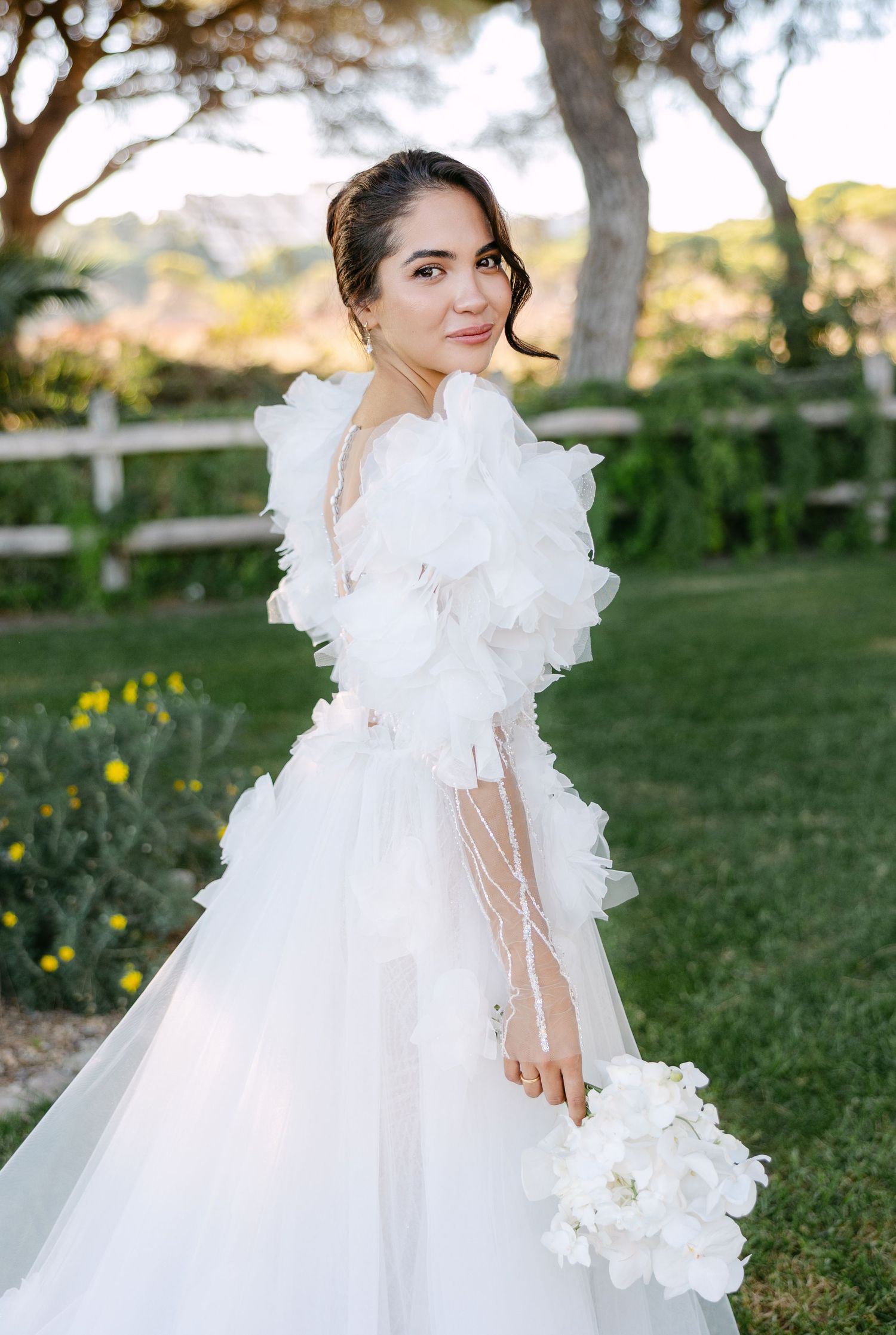 A bride in a romantic white gown with ruffled sleeves twirls in an outdoor garden setting.