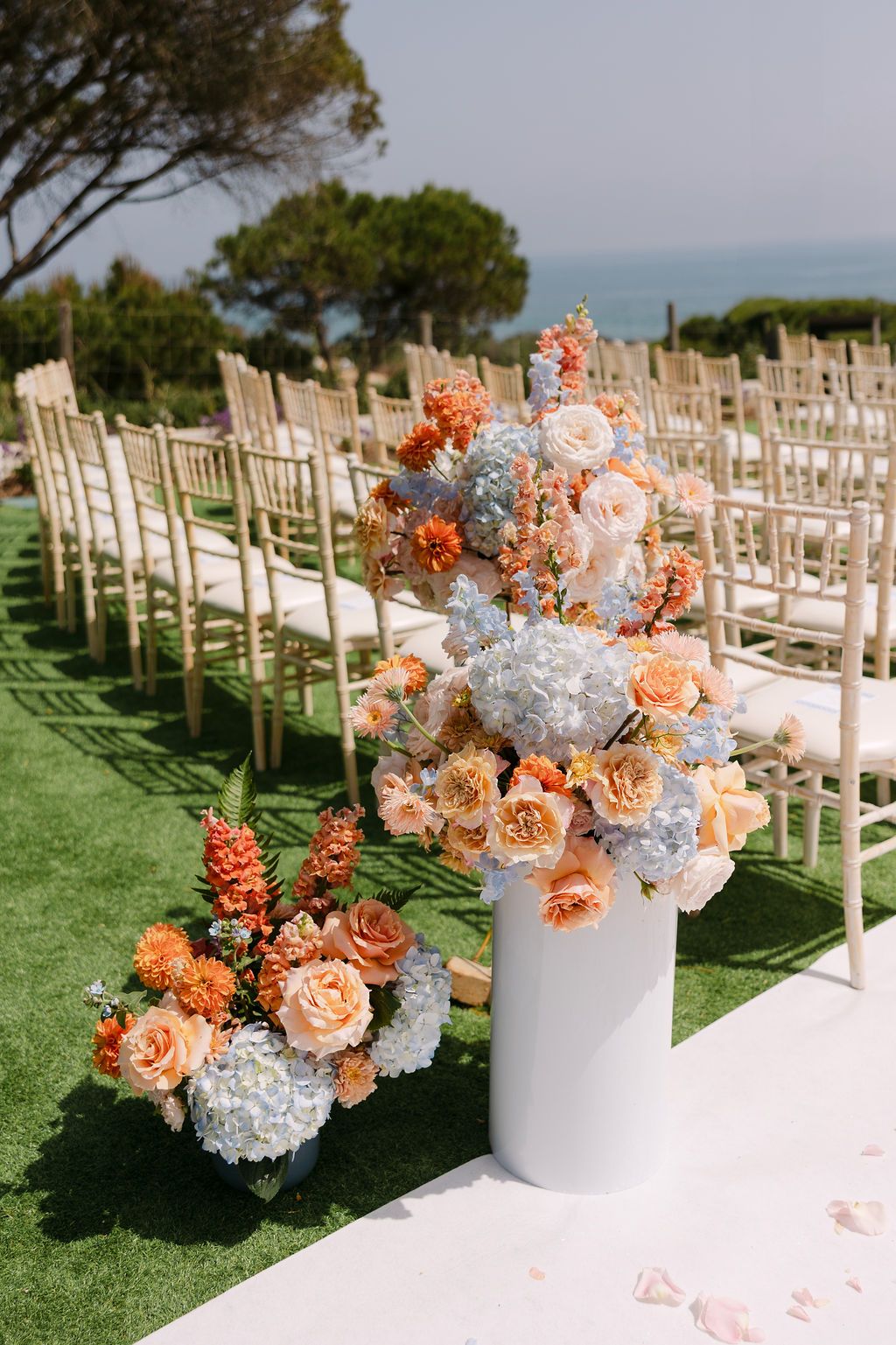 Outdoor wedding ceremony setup with white chairs and peach and blue floral arrangements along the aisle.