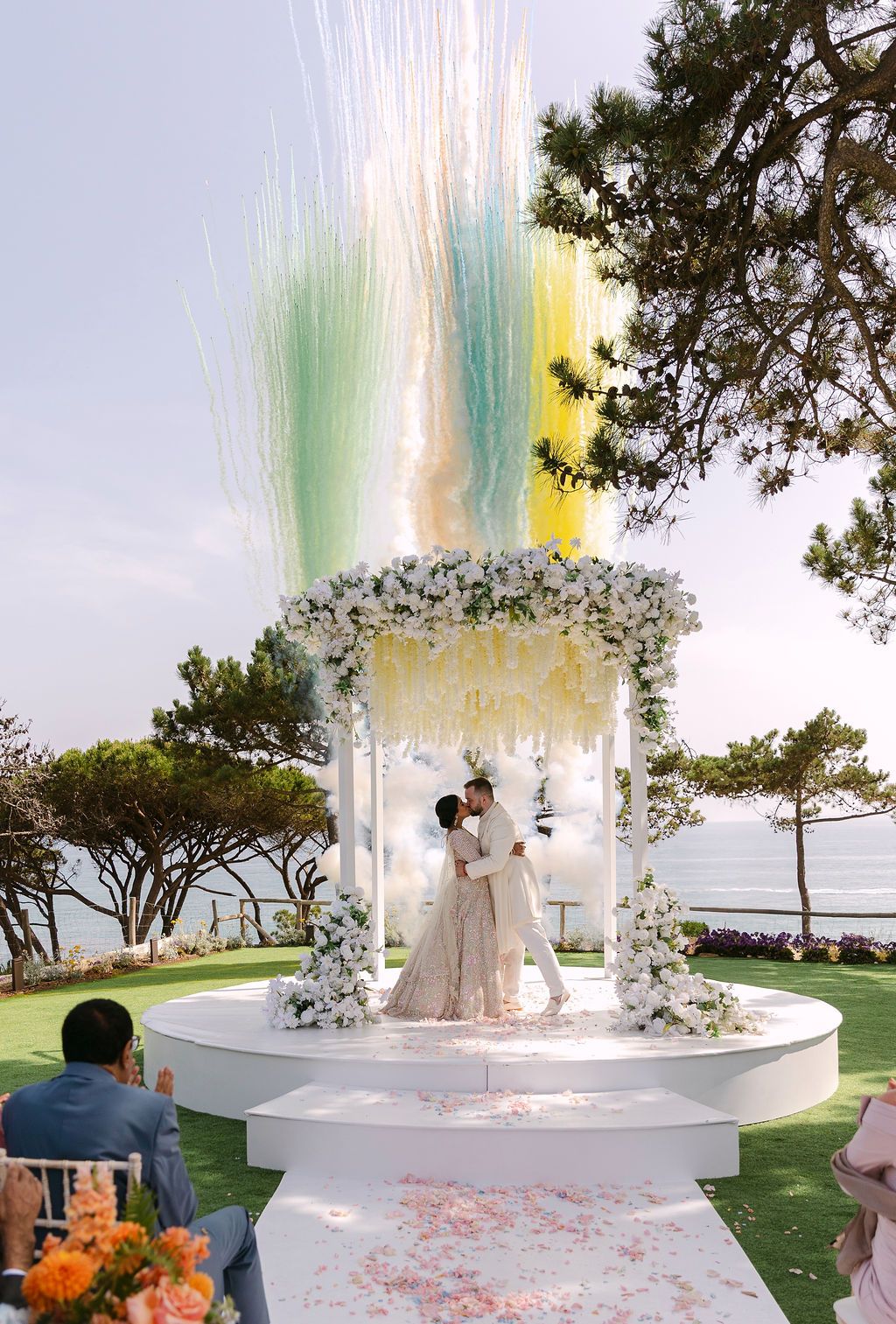 Wedding ceremony setup with a white floral arch and colorful smoke effects at sunset.