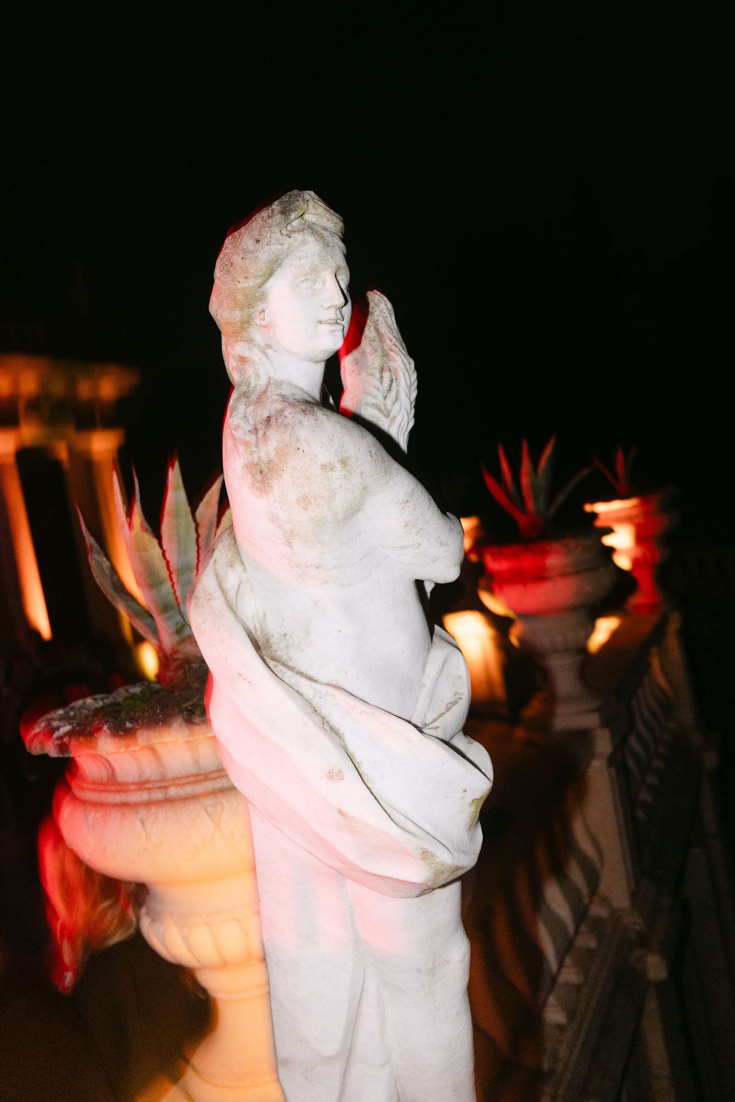 Illuminated classical stone statue at night with terracotta planters and warm ambient lighting.