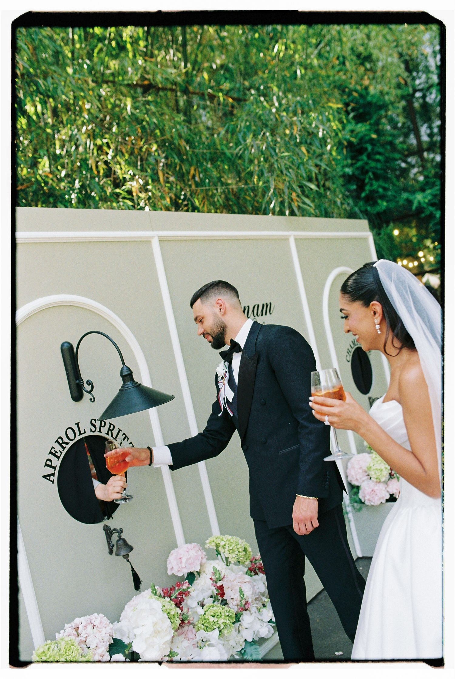 Bride and groom toasting with Aperol Spritz cocktails at an outdoor wedding reception near a floral display.
