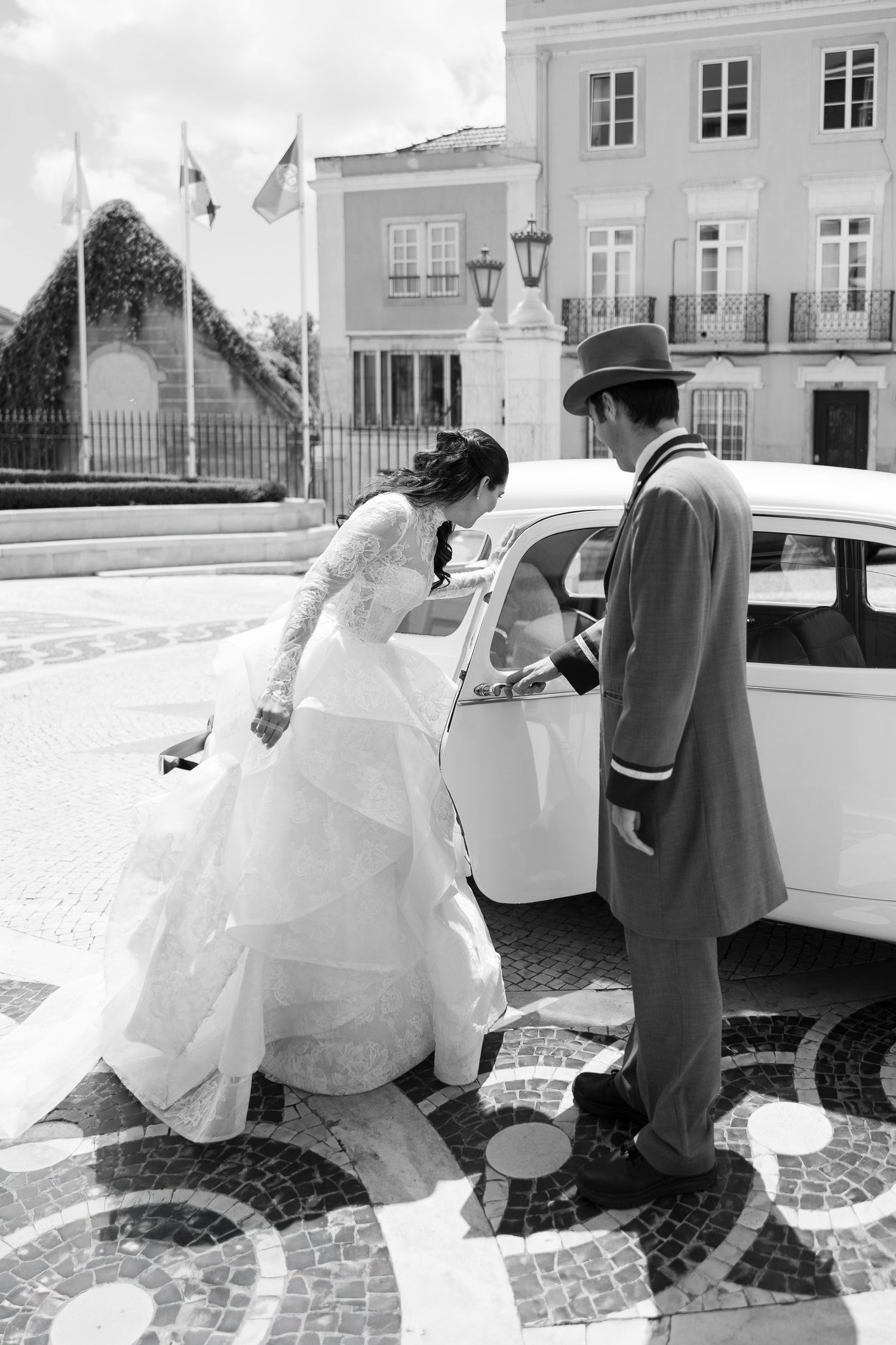 Classic black and white photo of bride entering vintage car on cobblestone street.