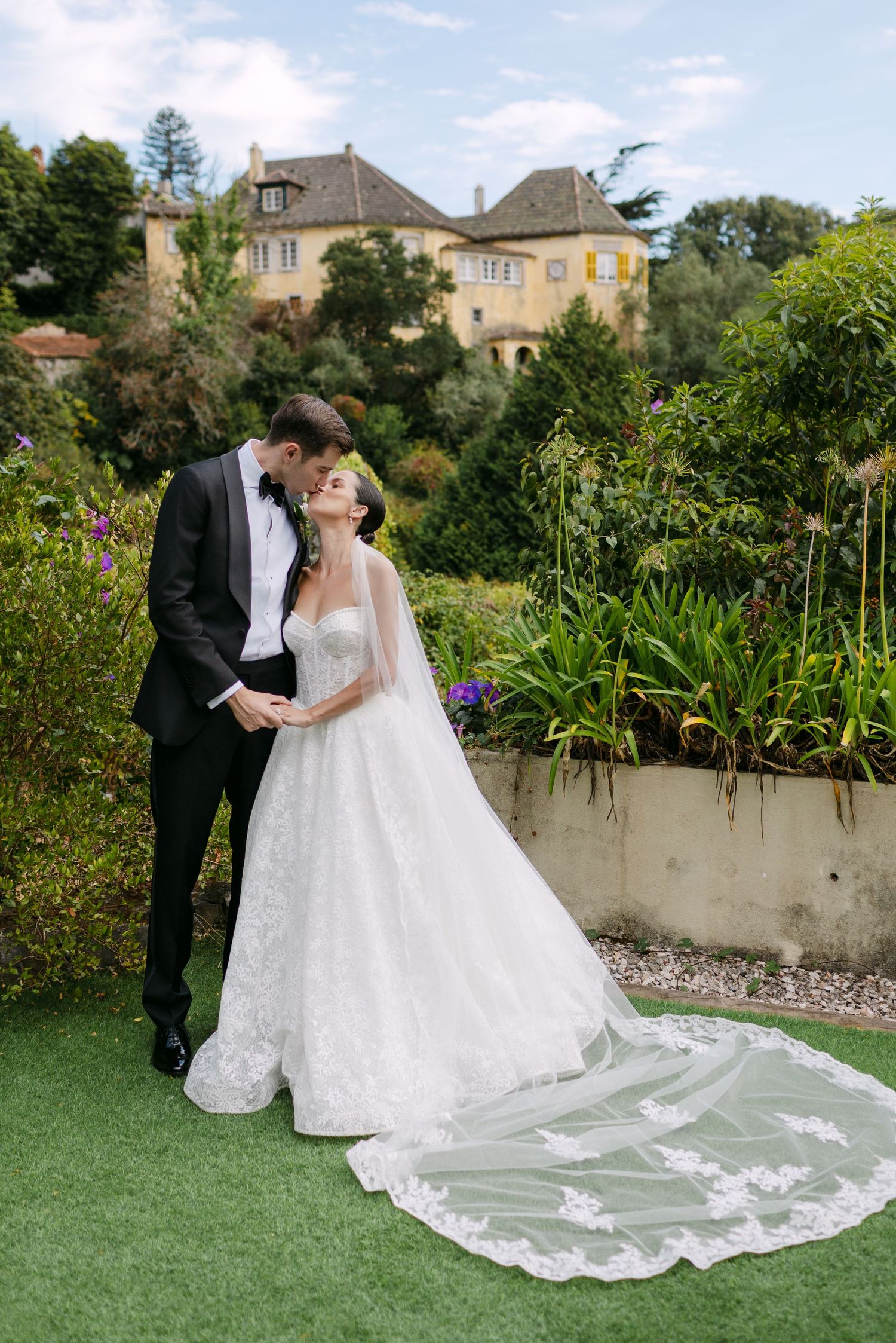 Wedding couple posing in a garden setting with a flowing lace train on the bride's dress.