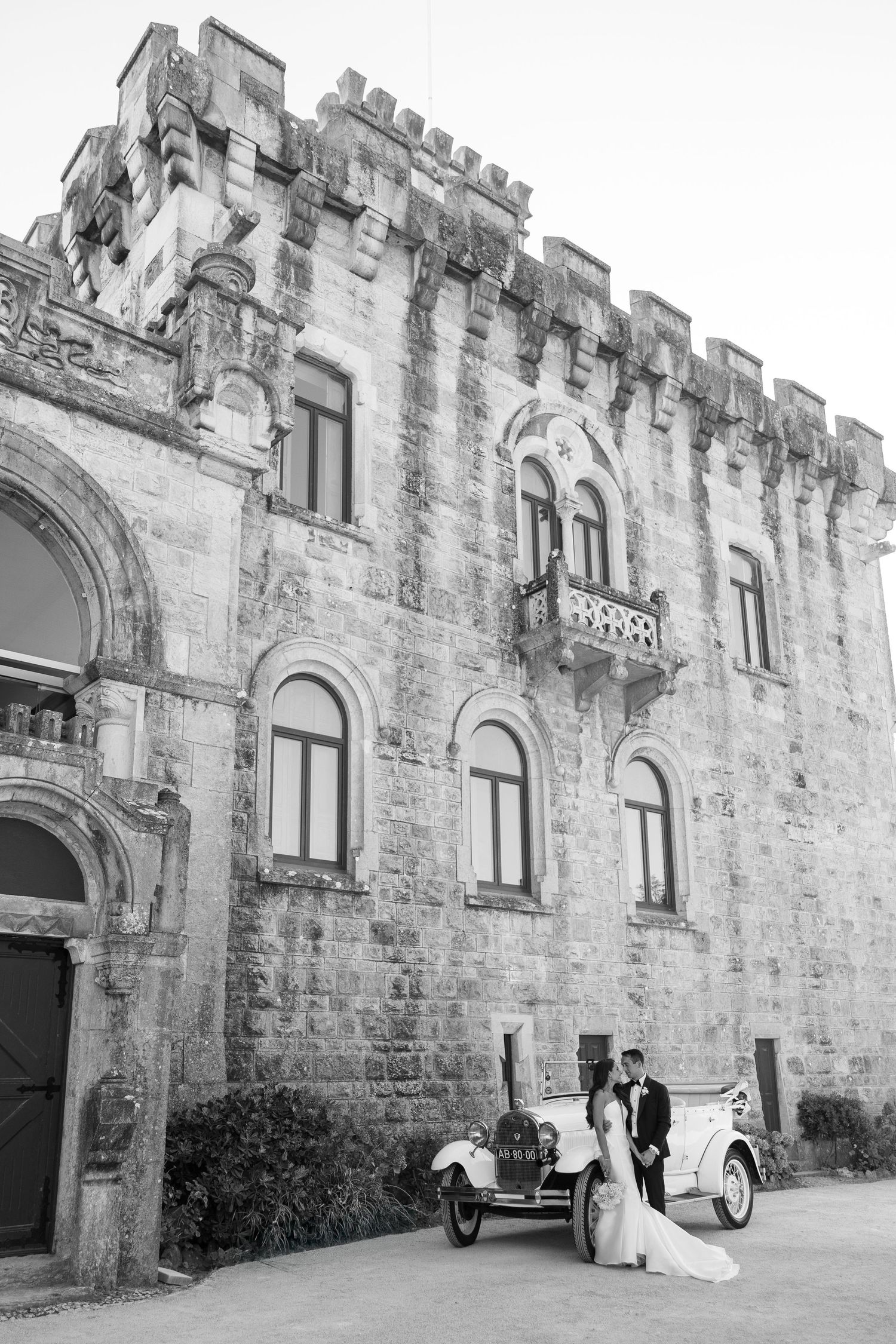 Wedding couple poses against historic stone castle exterior with arched windows.