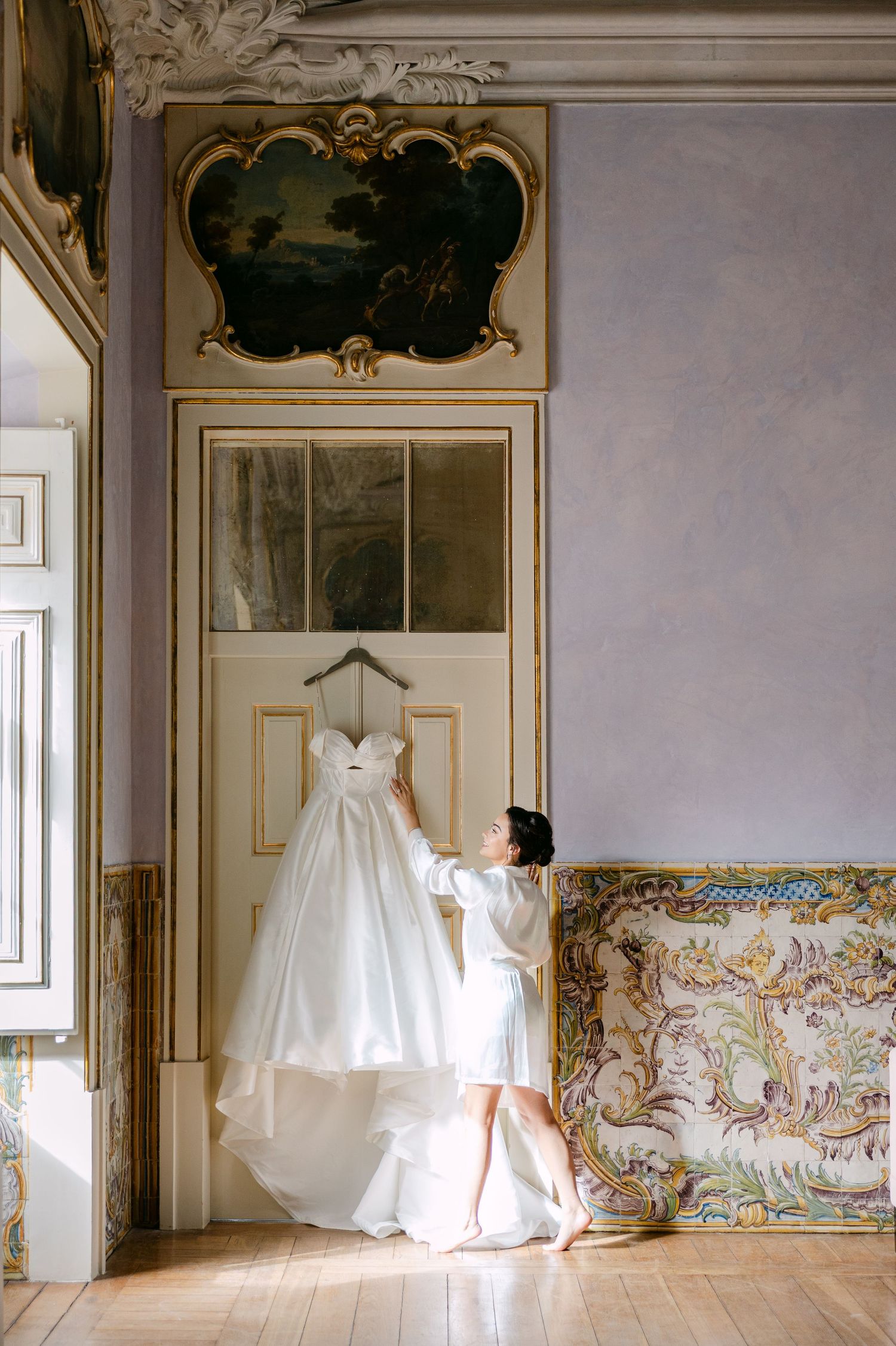 A white wedding dress hangs elegantly in an ornate room with decorative wall panels and gilded details.